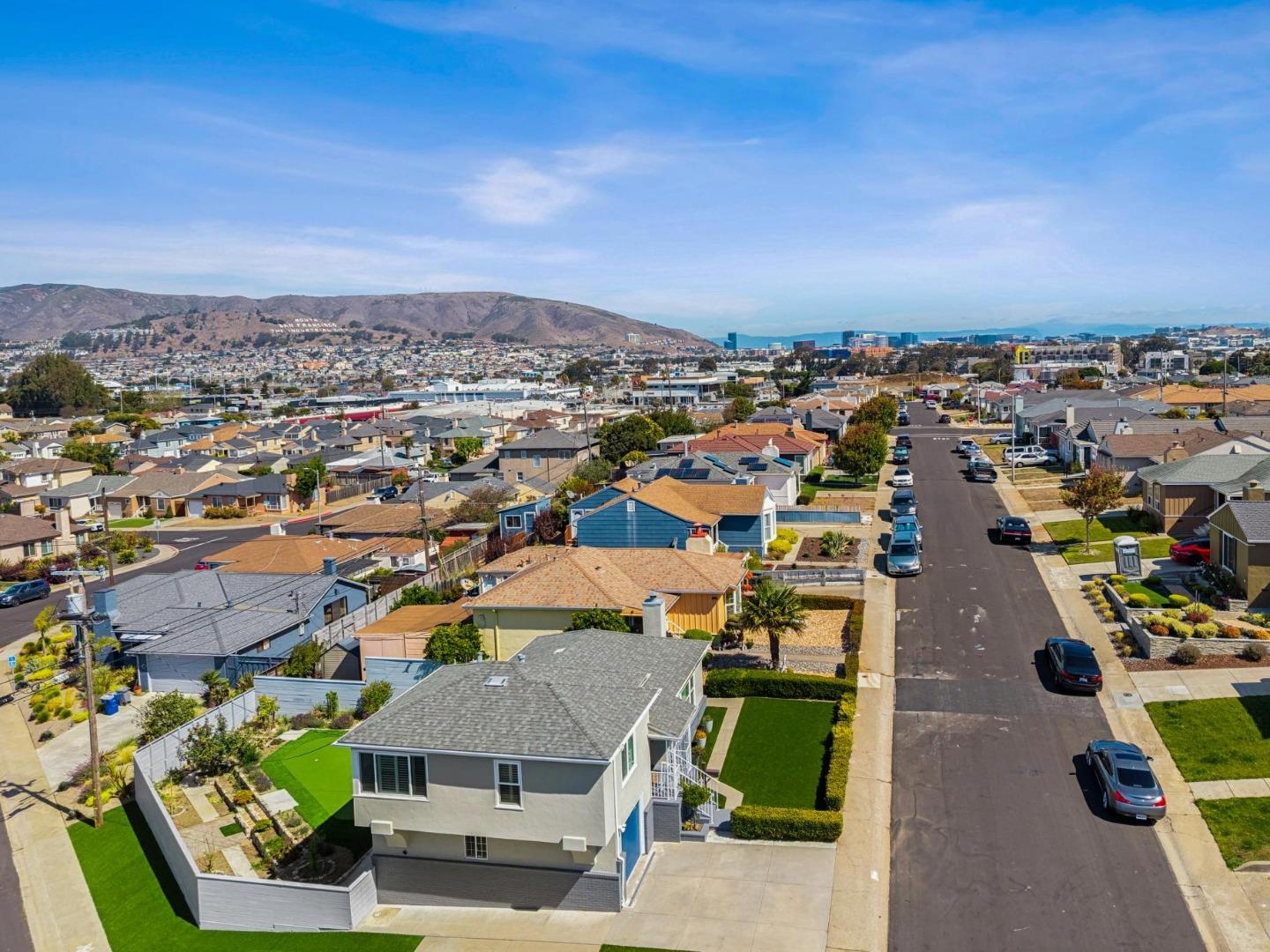 Aerial view of a two-story home and landscaped yard, with neighborhood streets, distant hills, and city buildings.
