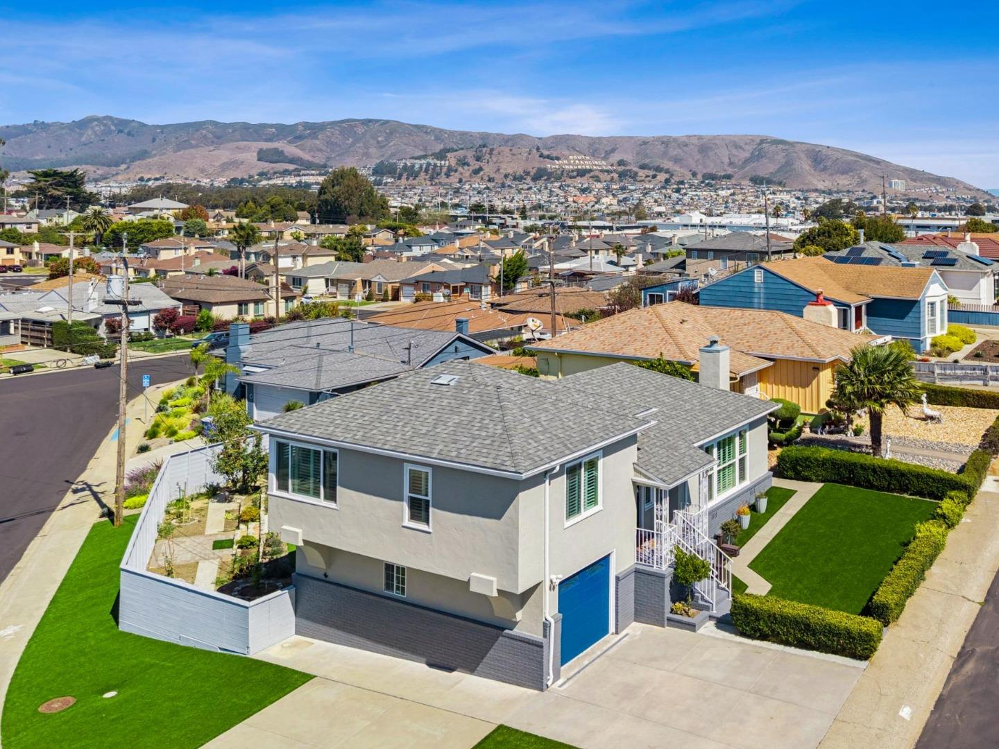 Aerial view of a gray two-story home with a blue garage door, green lawn, and South San Francisco hills beyond.