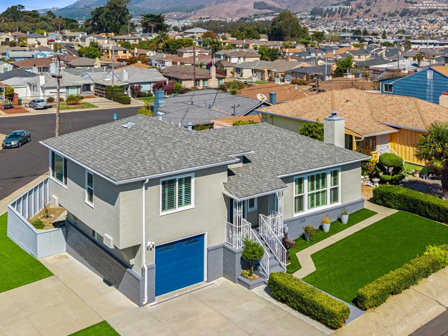 Gray two-story home with a blue garage door and landscaped lawn, viewed above the surrounding neighborhood.