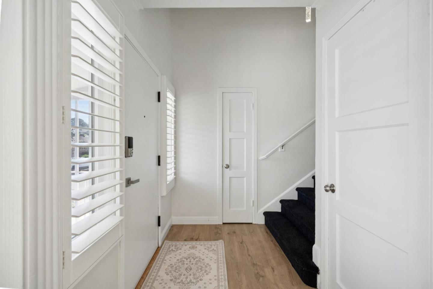 Bright entry hall with plantation shutters, a patterned runner, and dark carpeted stairs.
