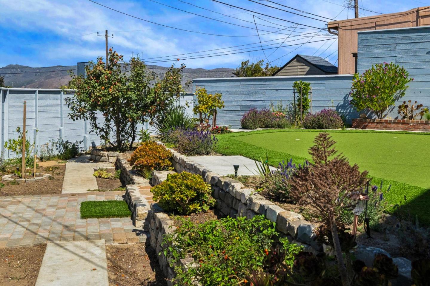 Backyard with a wide artificial turf lawn, stone-edged garden beds, and a paved patio.