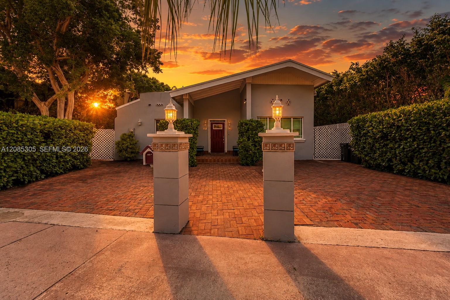 Modern house behind a brick courtyard, framed by tropical greenery beneath an orange sunset sky.