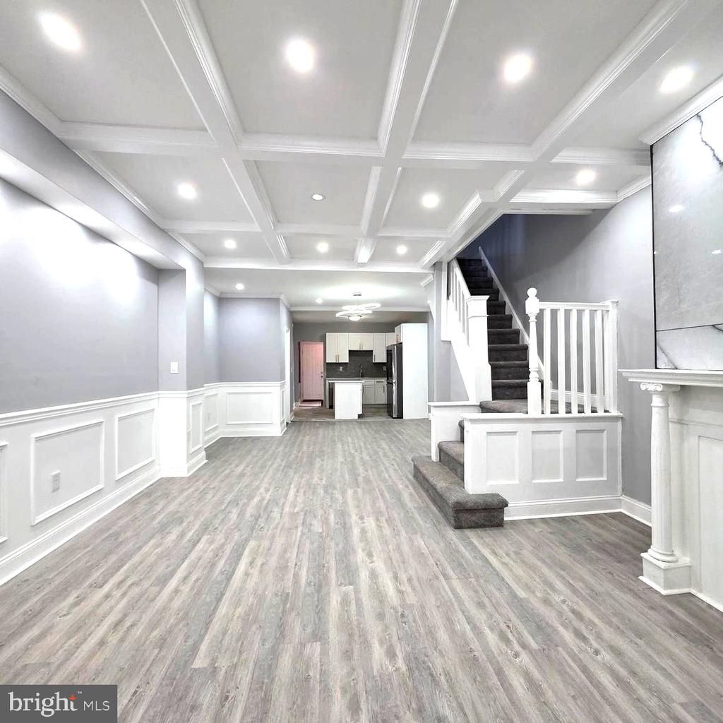 Unfurnished living room with gray plank flooring, white wainscoting, a coffered ceiling, and stairs beside the kitchen.