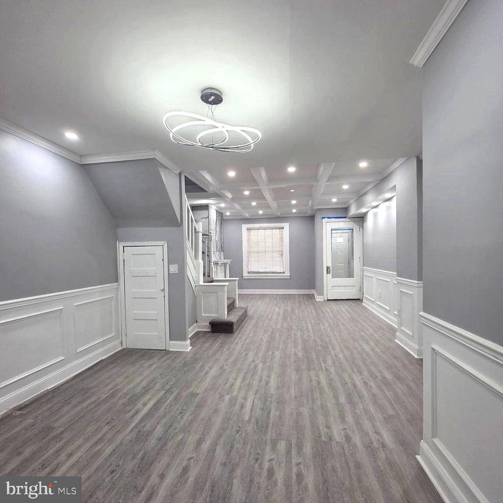 Empty renovated room with gray plank flooring, white wainscoting, staircase, and modern ring chandelier.