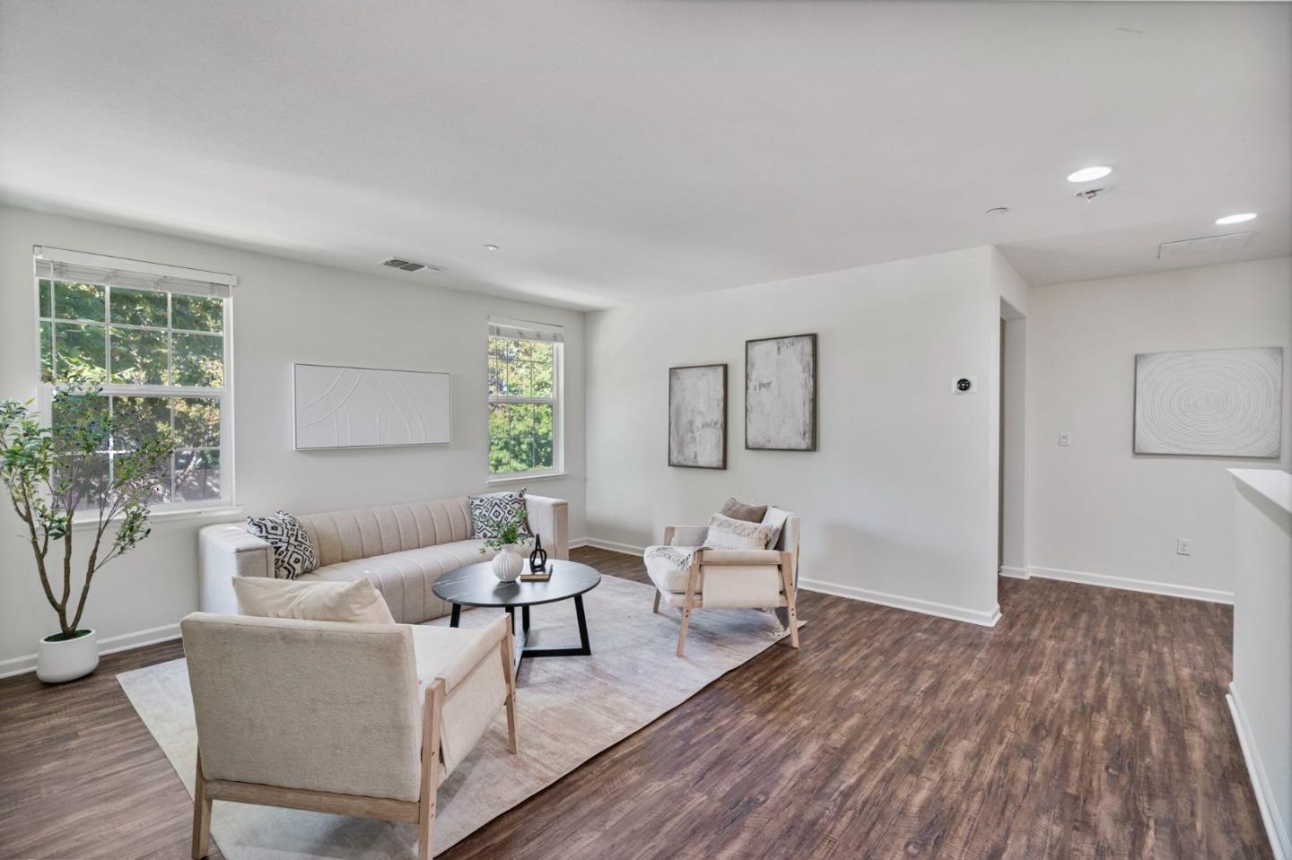 Staged living room with a light sofa, armchairs, large windows, and dark wood-look floors.