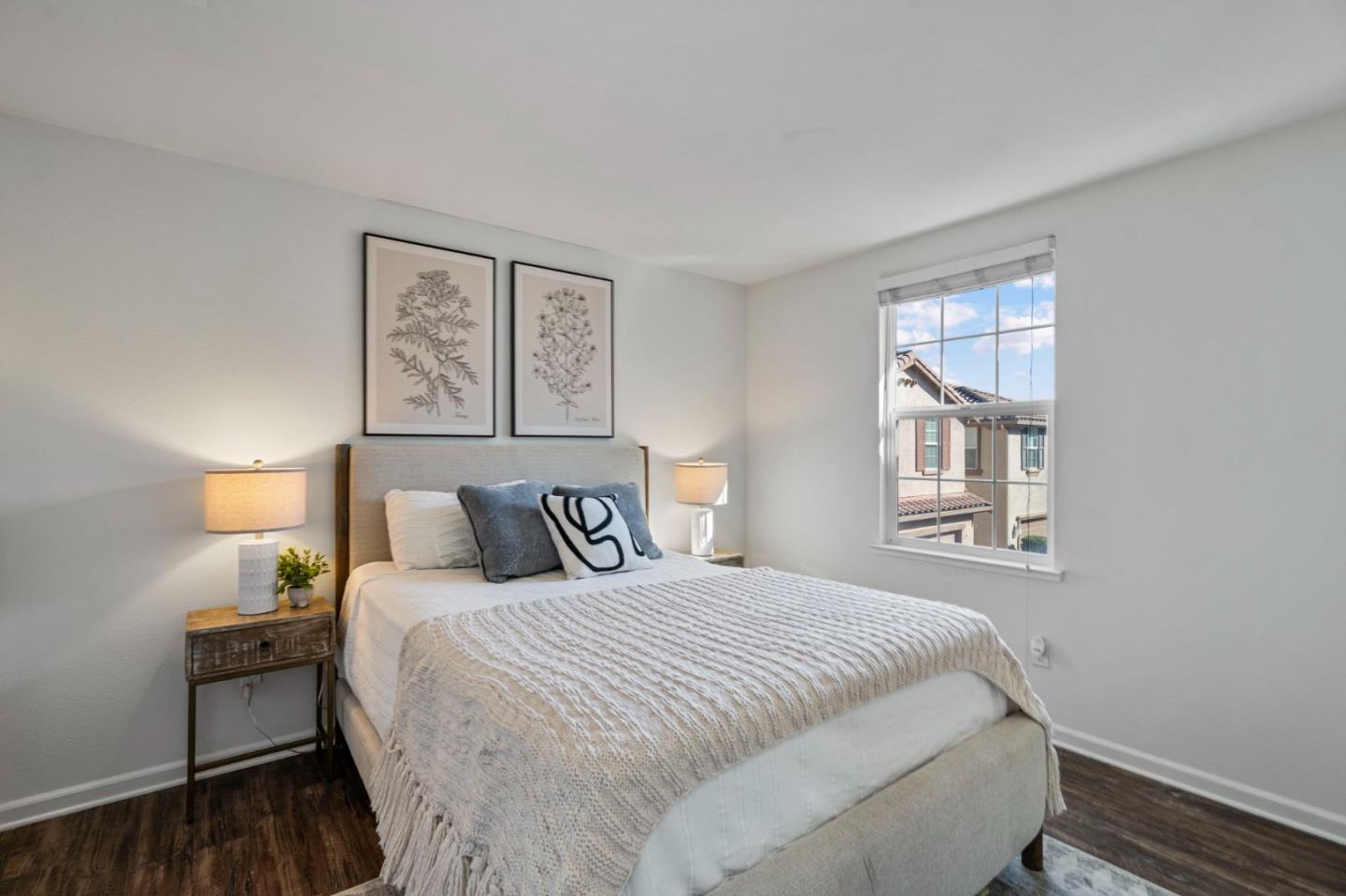 Staged bedroom with a white-and-gray bed, botanical prints, bedside lamps, and a window overlooking a neighboring home.