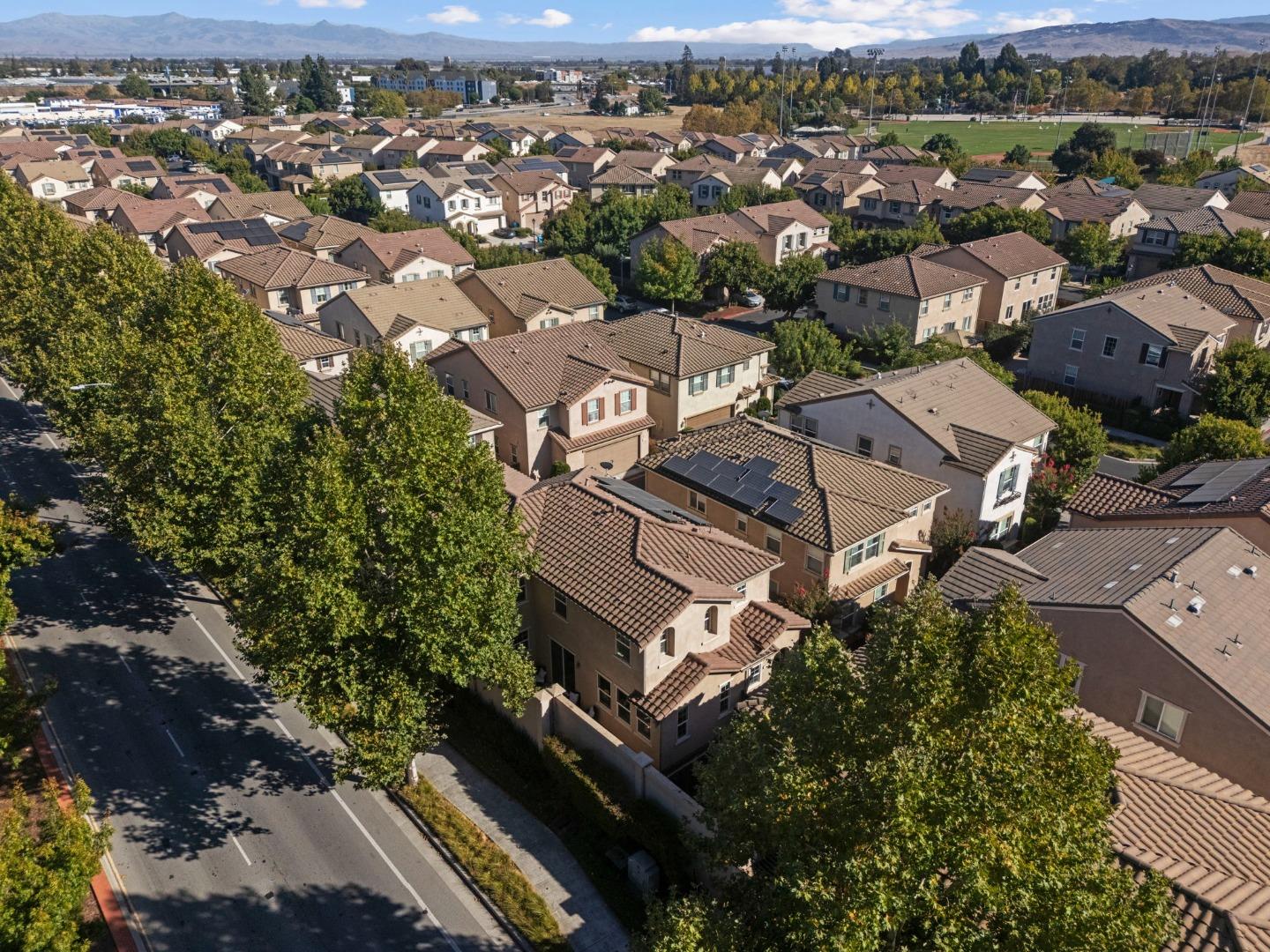 Aerial view of tiled-roof homes among mature trees, with sports fields and mountains in the distance.