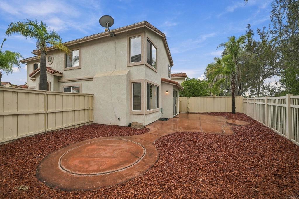 Two-story stucco home beside a fenced backyard with reddish mulch and a curved concrete patio.