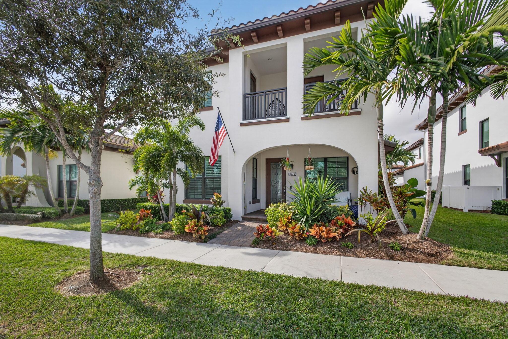 Two-story white stucco home with an arched entrance, balconies, tropical landscaping, and an American flag.