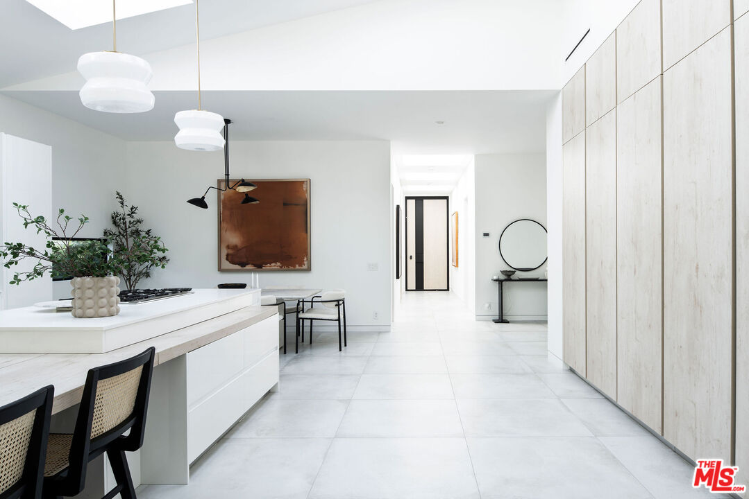 Modern white kitchen with a large island, dining table, pendant lights, and pale stone floors.