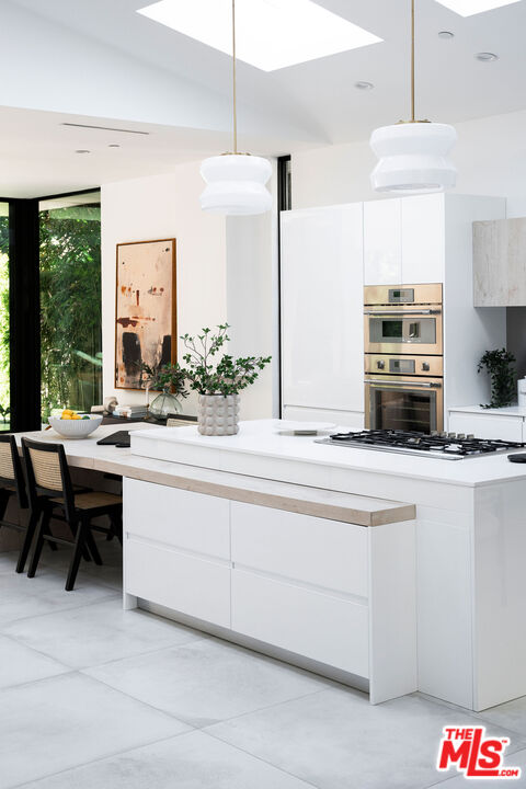 Modern white kitchen with a large island, built-in ovens, skylights, and windows overlooking greenery.