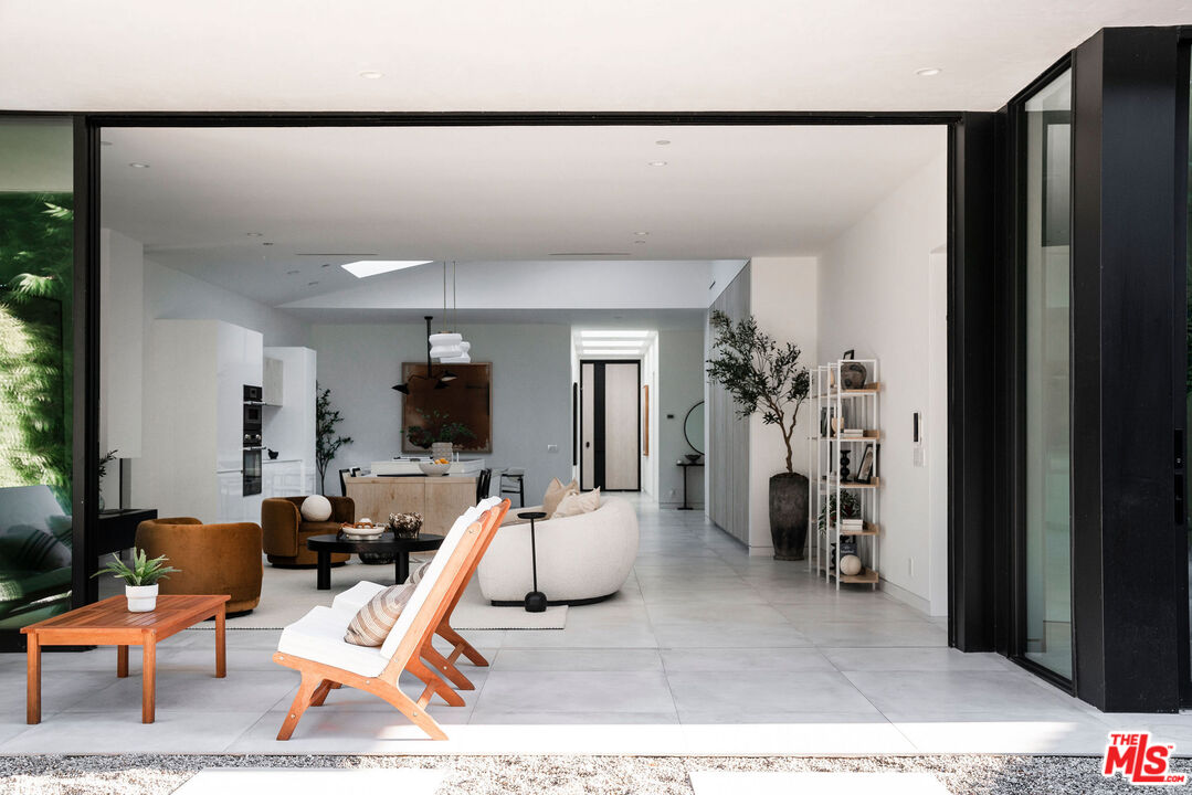 Open-plan living room and kitchen with pale tile floors, sculptural seating, and wide-open sliding glass doors.
