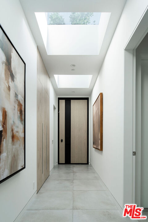 Modern hallway with two skylights, gray tile floors, abstract art, and a wood-and-black door.