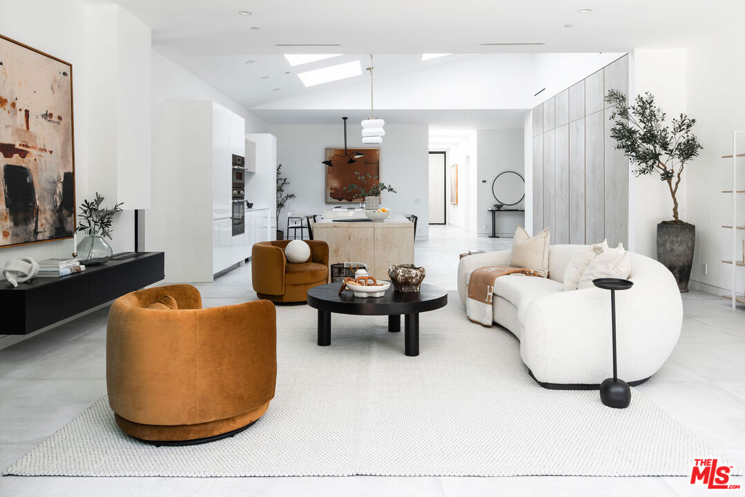 Open-plan living room with a curved white sofa, rust-colored chairs, and light wood kitchen cabinetry.