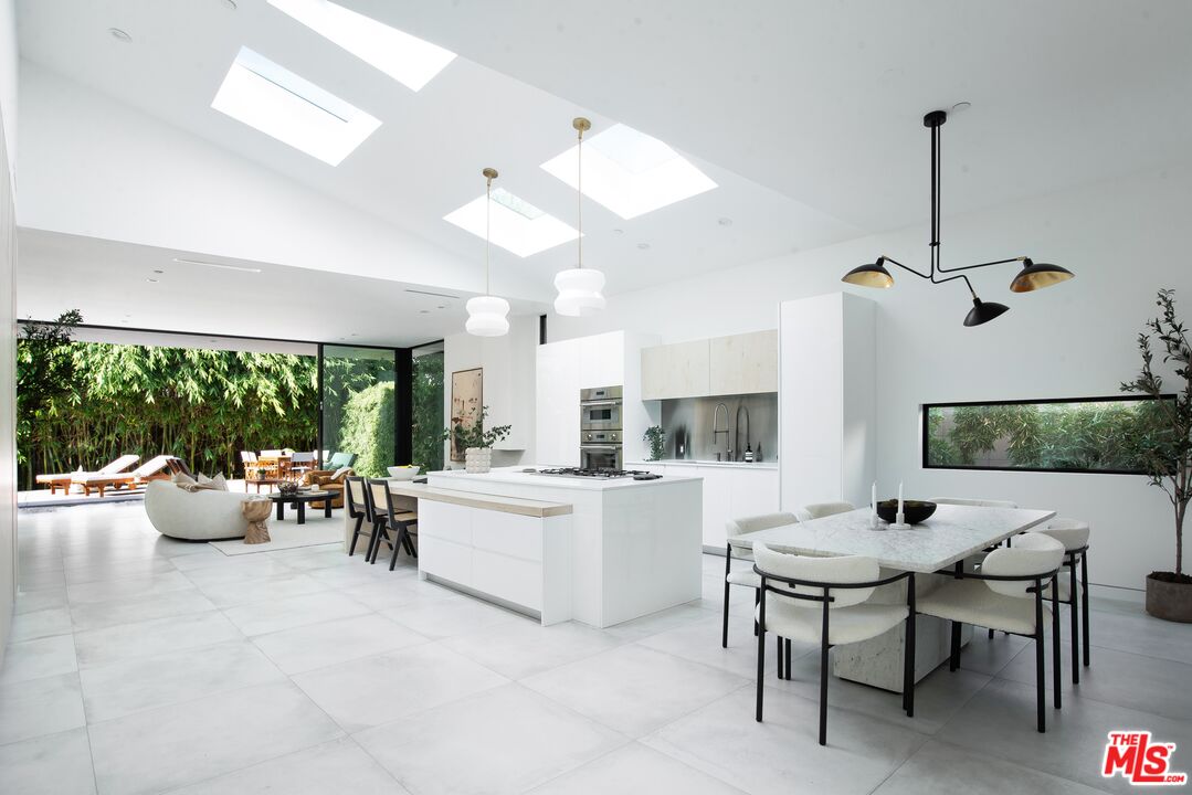 Modern white kitchen with a large island, skylights, and dining area beside floor-to-ceiling glass.