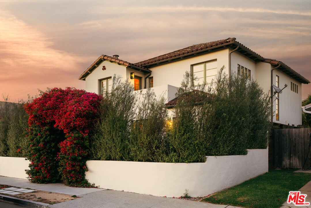 Two-story stucco home with a red tile roof, tall hedges, and a red-flowering shrub at sunset.