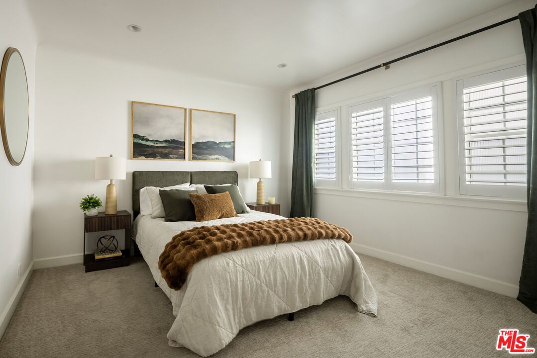 Bright staged bedroom with a white bed, dark headboard, and plantation shutters.