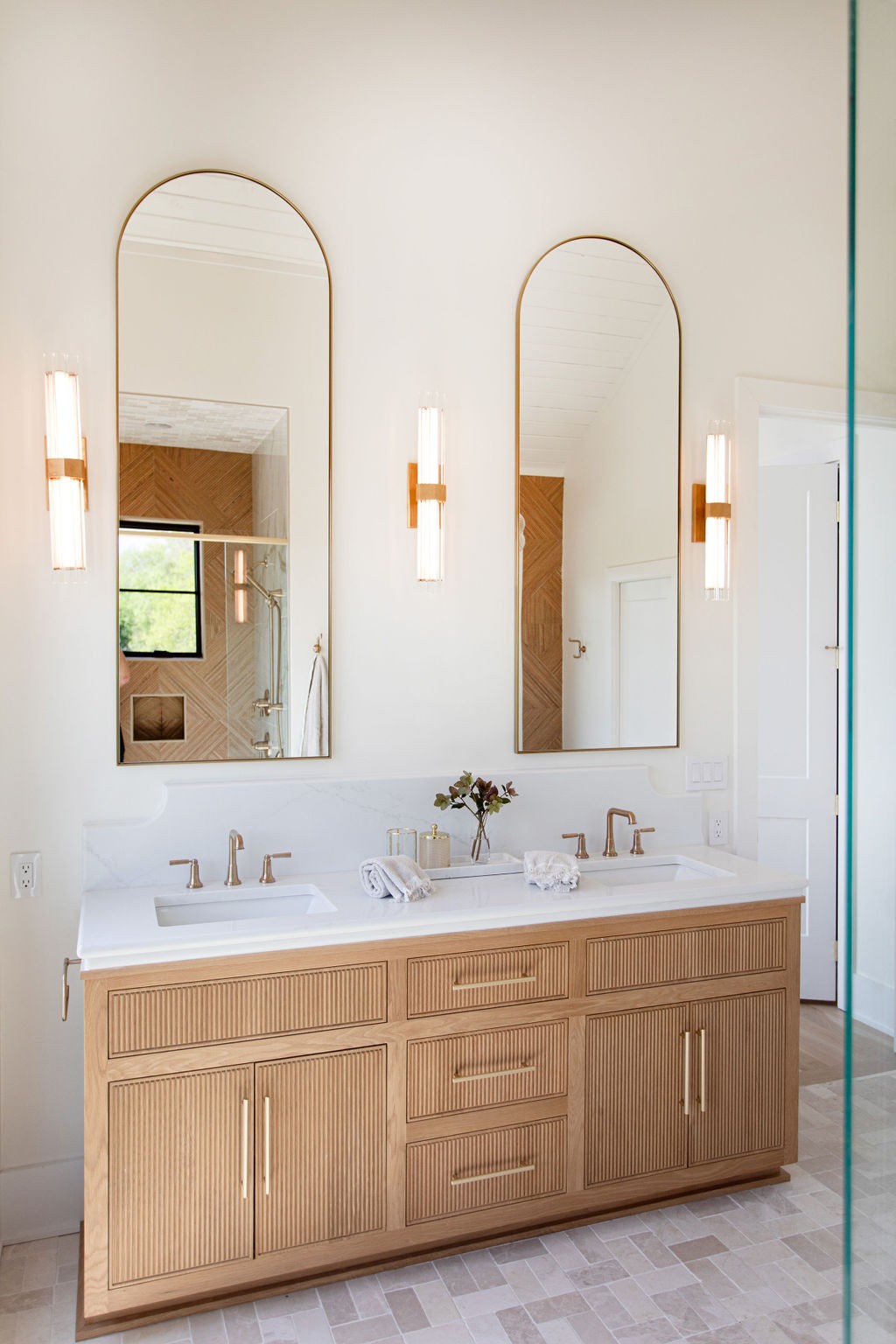 Bathroom with a fluted wood double vanity, brass fixtures, and two tall arched mirrors.