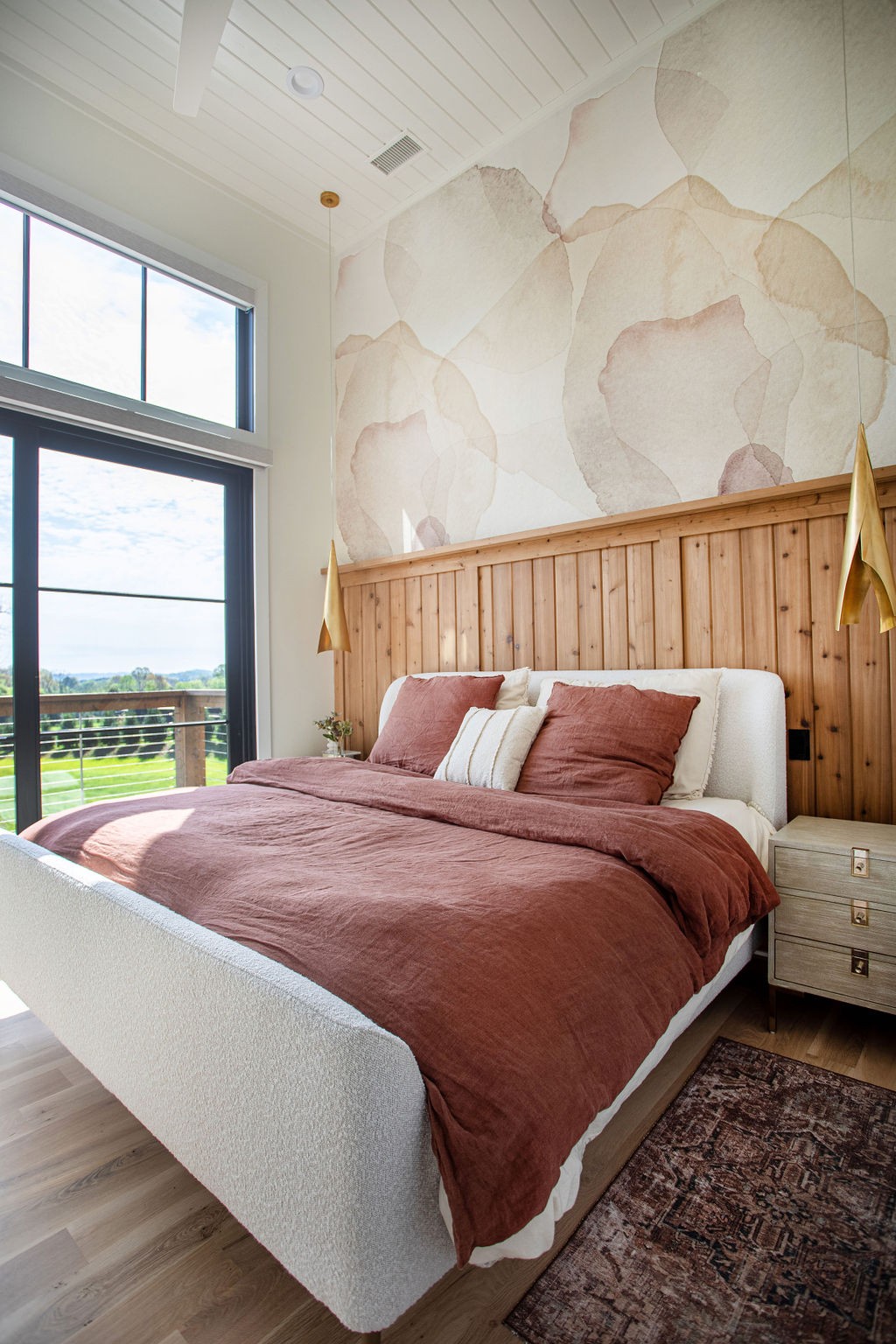 Bedroom with rose bedding, a wood-paneled headboard wall, and tall windows overlooking greenery.