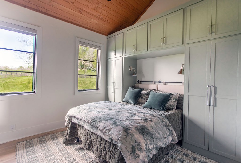 Bedroom with a king-size bed, built-in pale green cabinetry, vaulted wood ceiling, and windows overlooking a fenced lawn.