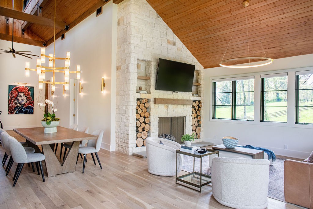 Open-plan living and dining room with a stone fireplace, vaulted wood ceiling, and windows overlooking trees.