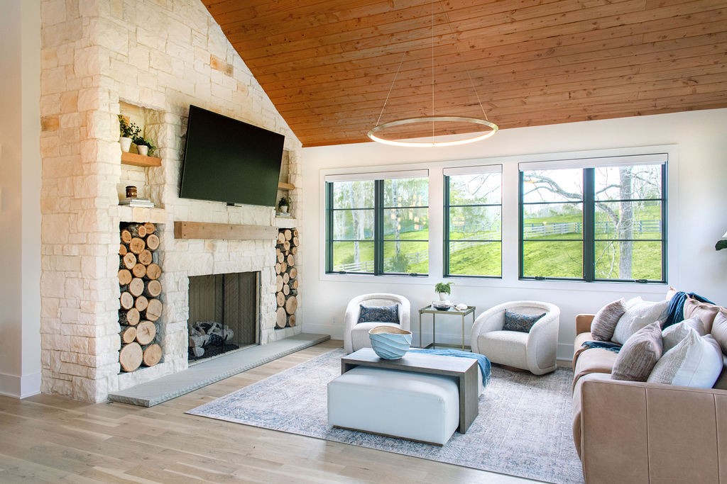 Living room with a stone fireplace, vaulted wood ceiling, and large windows overlooking a green yard.