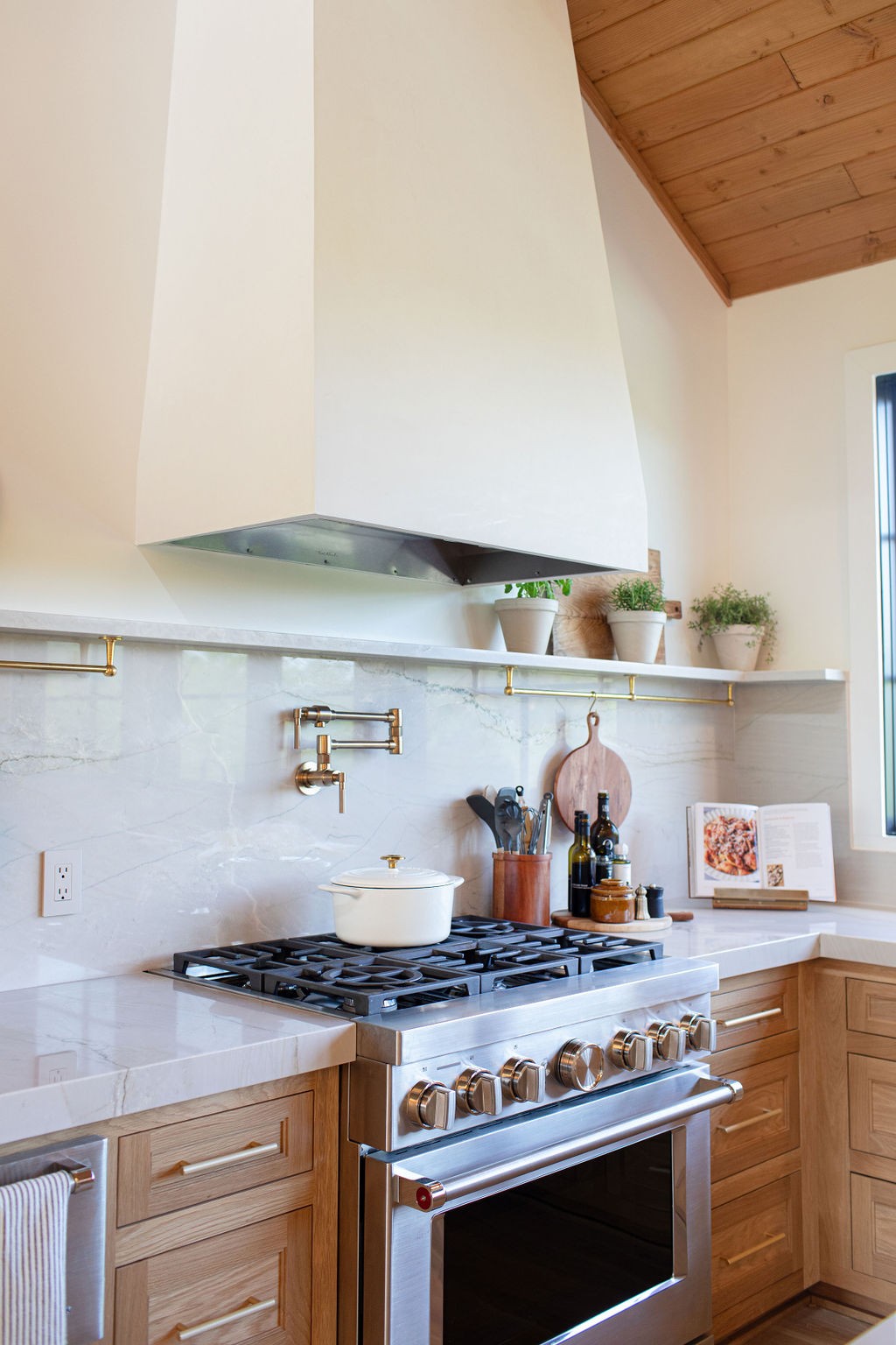 Kitchen with a stainless gas range, oversized white hood, marble backsplash, and warm wood cabinetry.