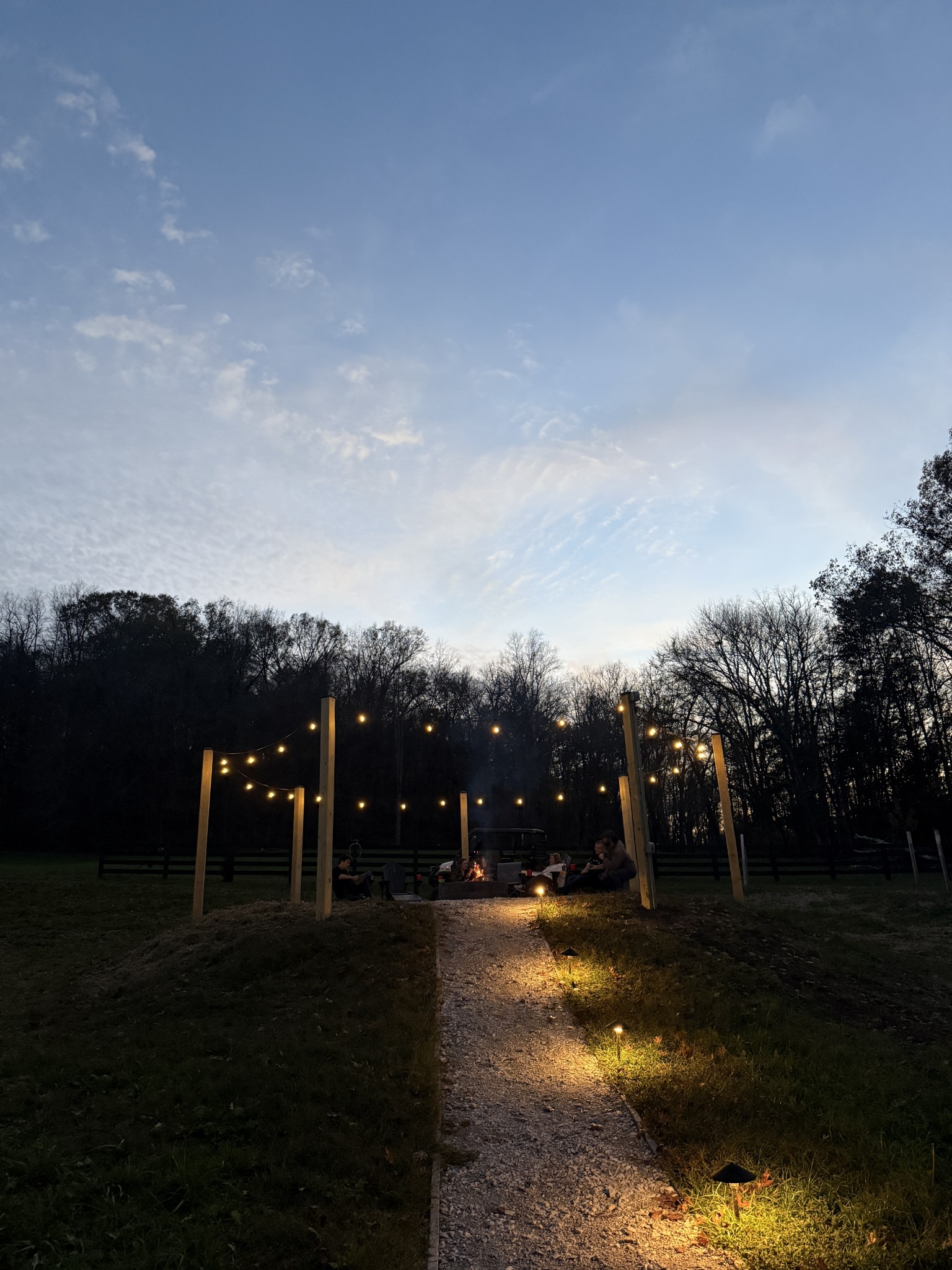 Backyard fire pit with string lights, people gathered around the fire, and a lit gravel path.
