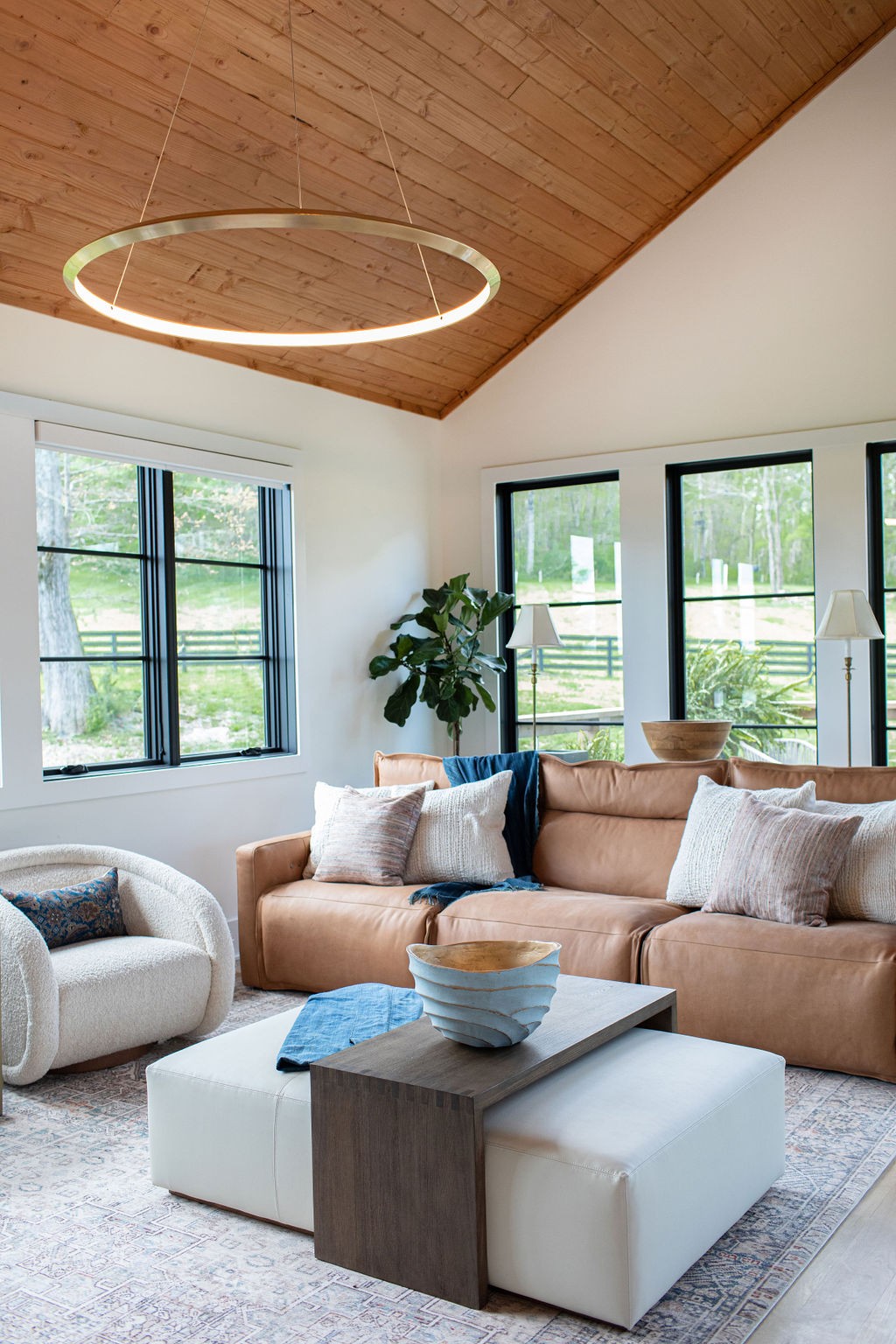 Living room with a vaulted wood ceiling, circular pendant light, and large windows overlooking a green yard.