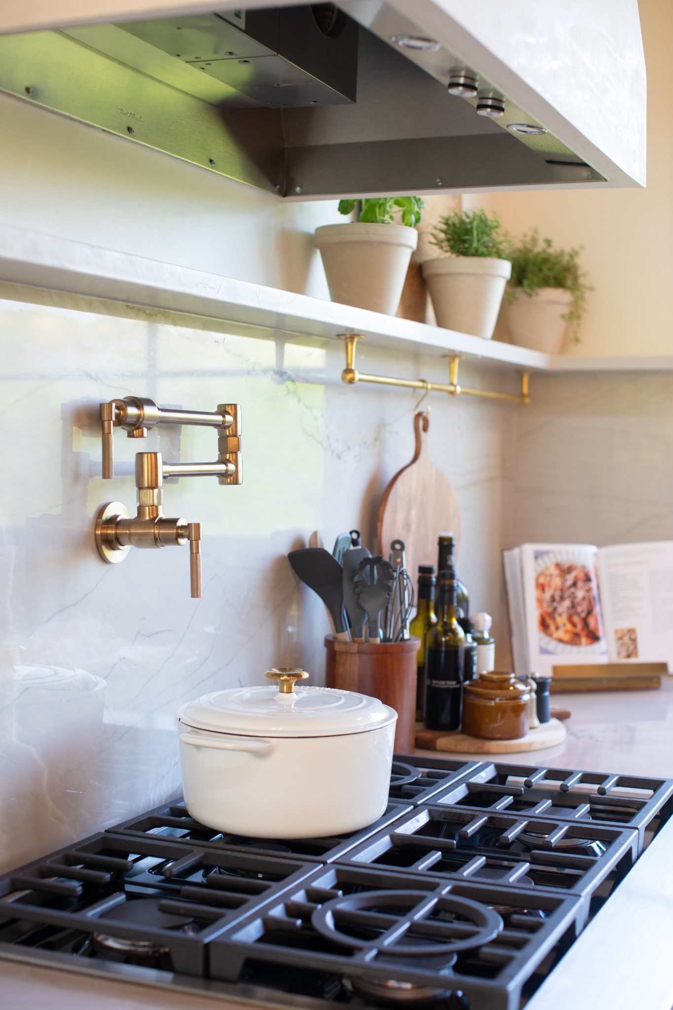 Kitchen cooktop with a white pot, brass pot filler, marble backsplash, and potted herbs.