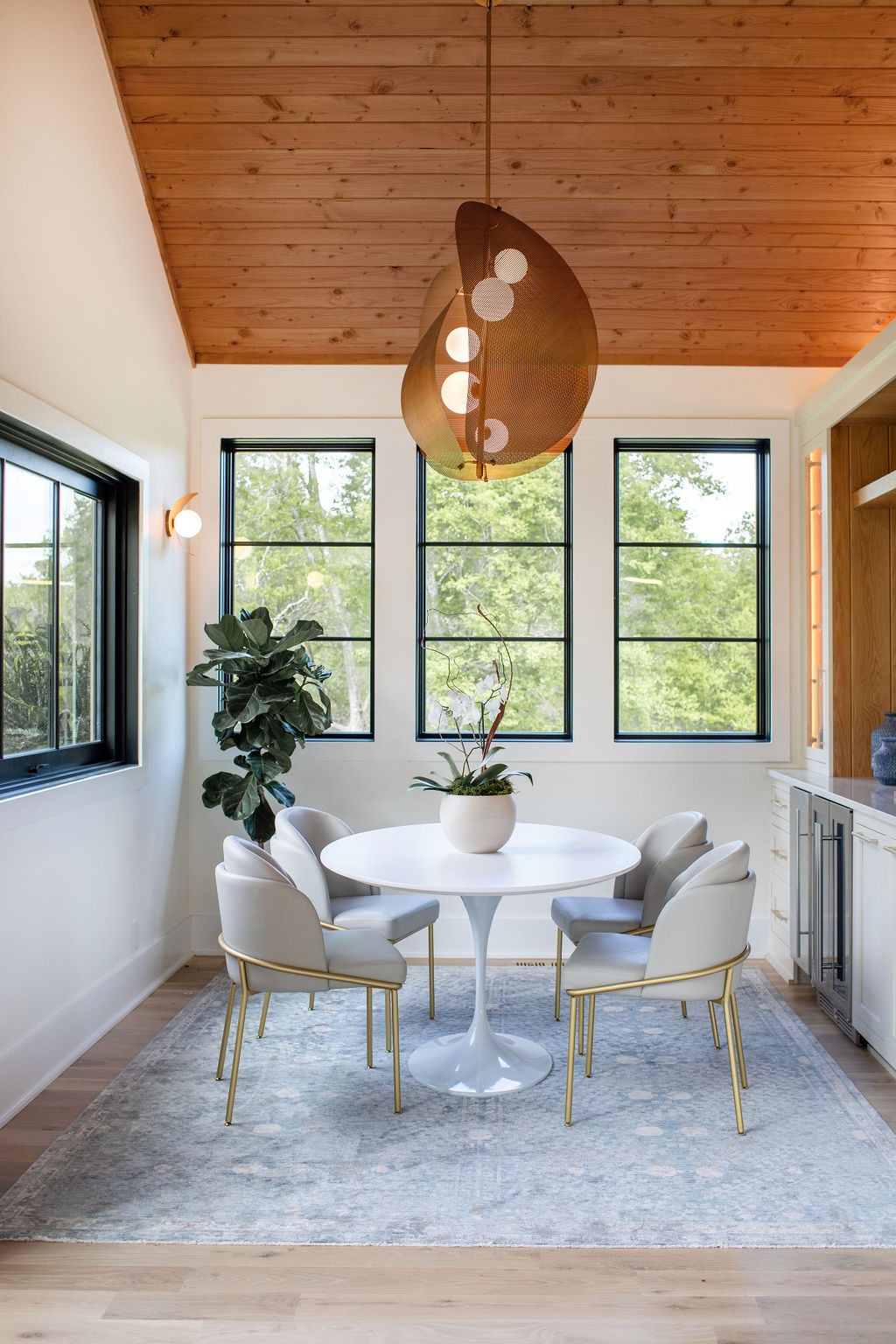 Dining area with a round pedestal table, sculptural pendant light, and wood-plank ceiling.
