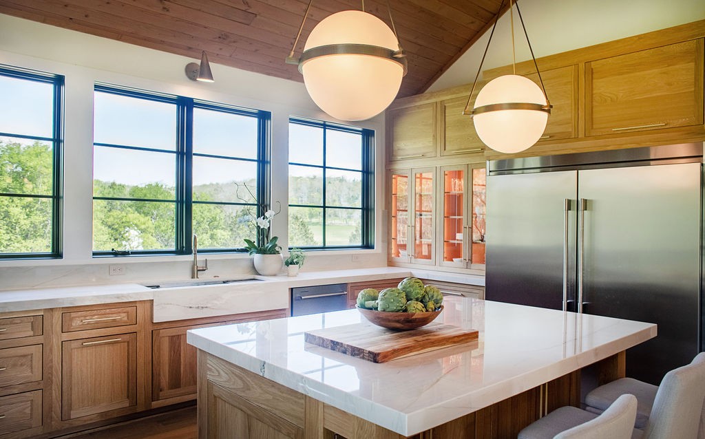 Bright kitchen with a marble island, wood cabinetry, and large windows overlooking trees.