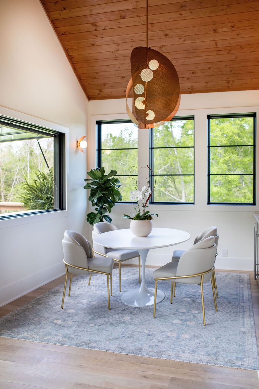 Dining area with a round white table, four chairs, large windows, and a vaulted wood ceiling.
