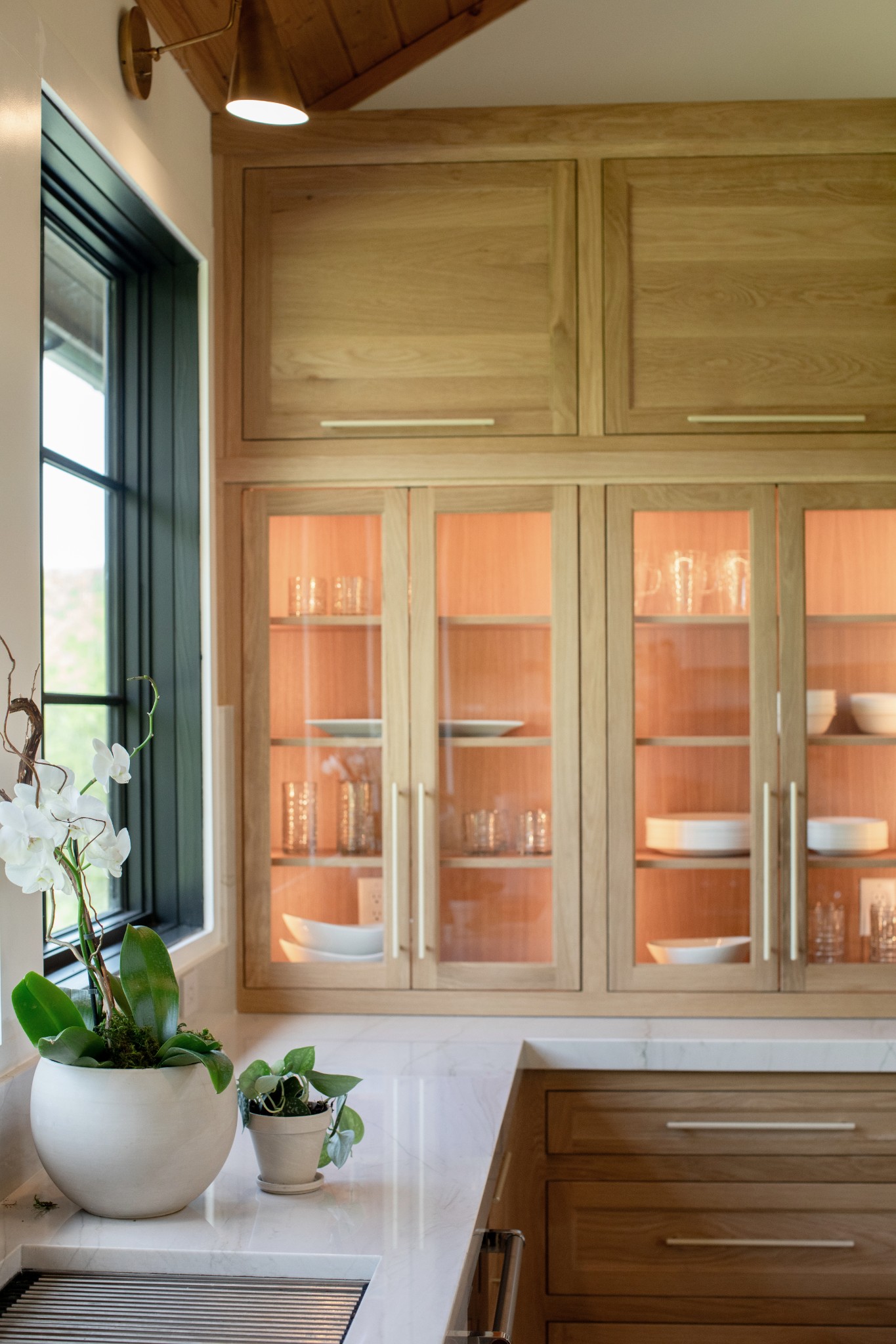 Kitchen with glass-front wood cabinets, a marble countertop, and orchids by a black-framed window.