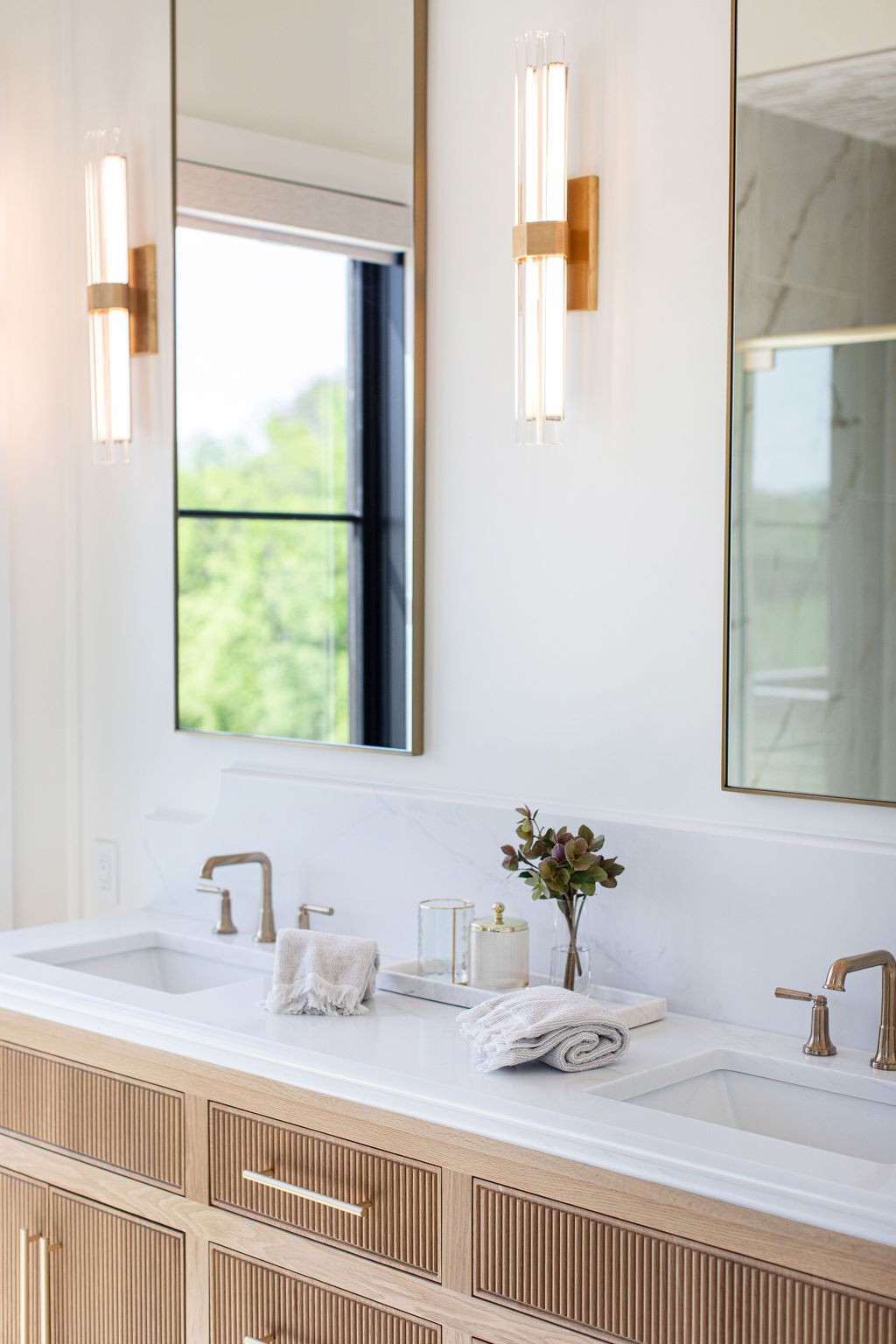 Bathroom vanity with two sinks, fluted light-wood cabinetry, brass faucets, and tall mirrors.