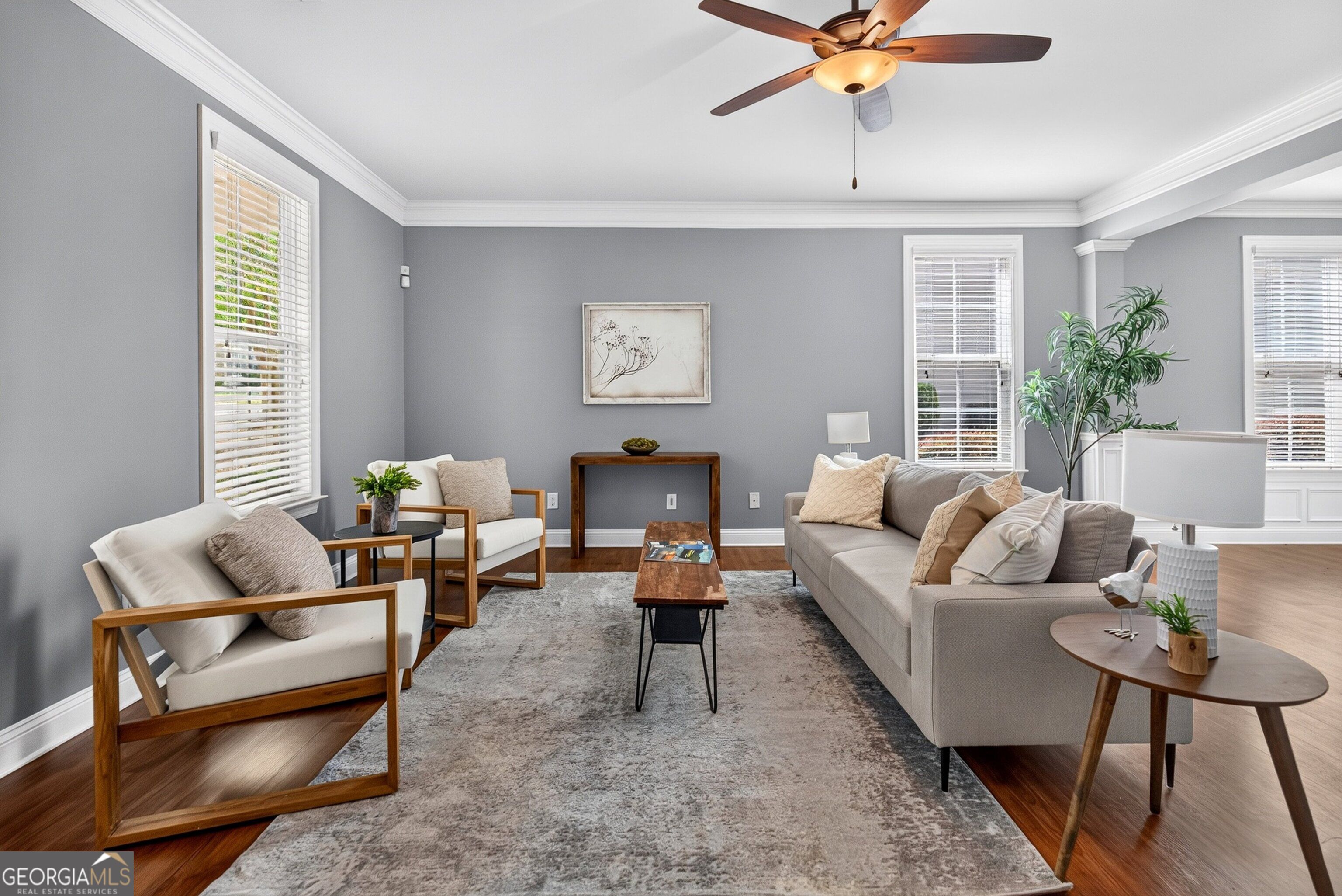 Staged living room with a gray sofa, white armchairs, hardwood floors, and large windows.