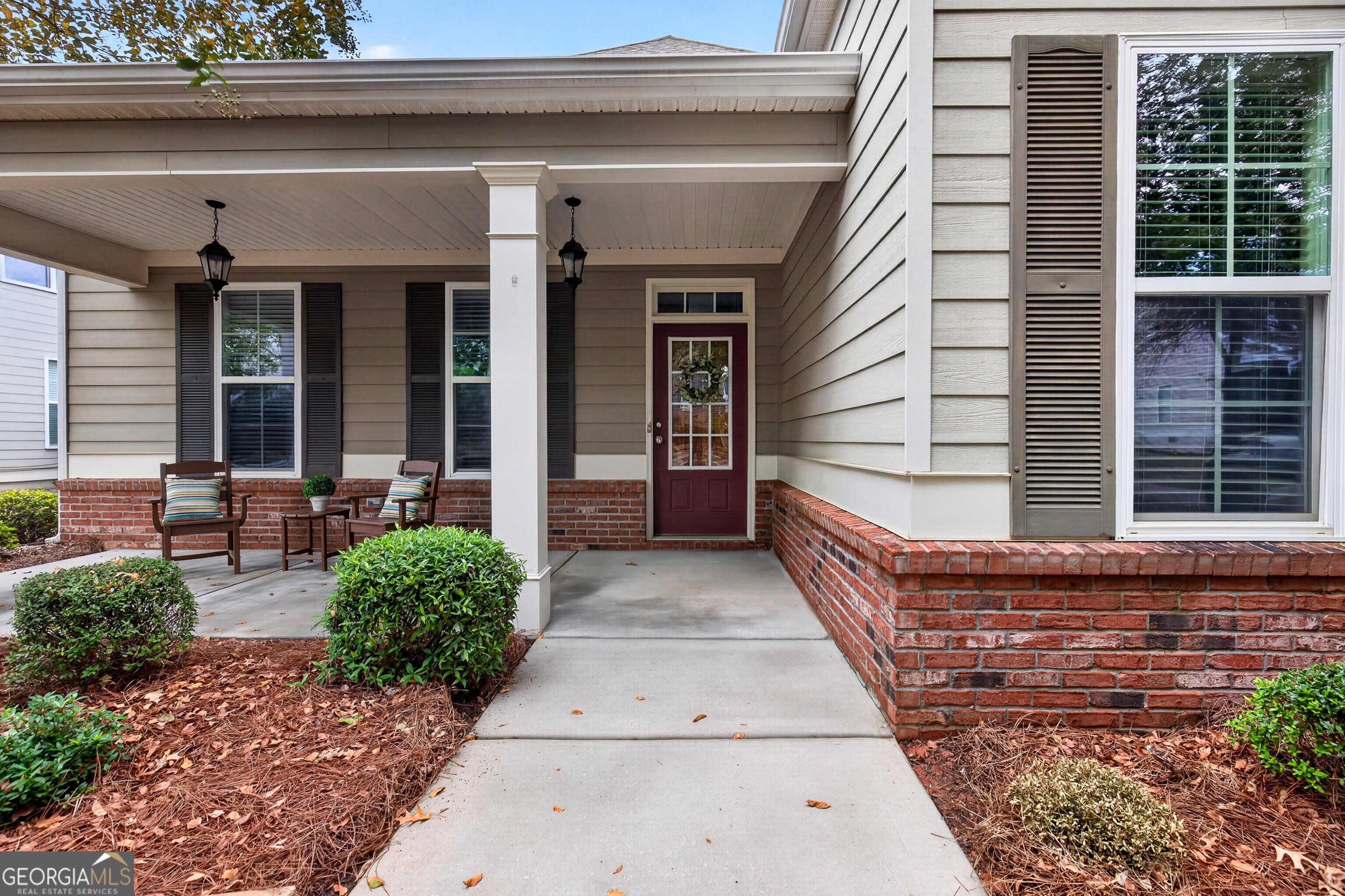House front porch with a burgundy door, white columns, brick foundation, and seating beside the walkway.