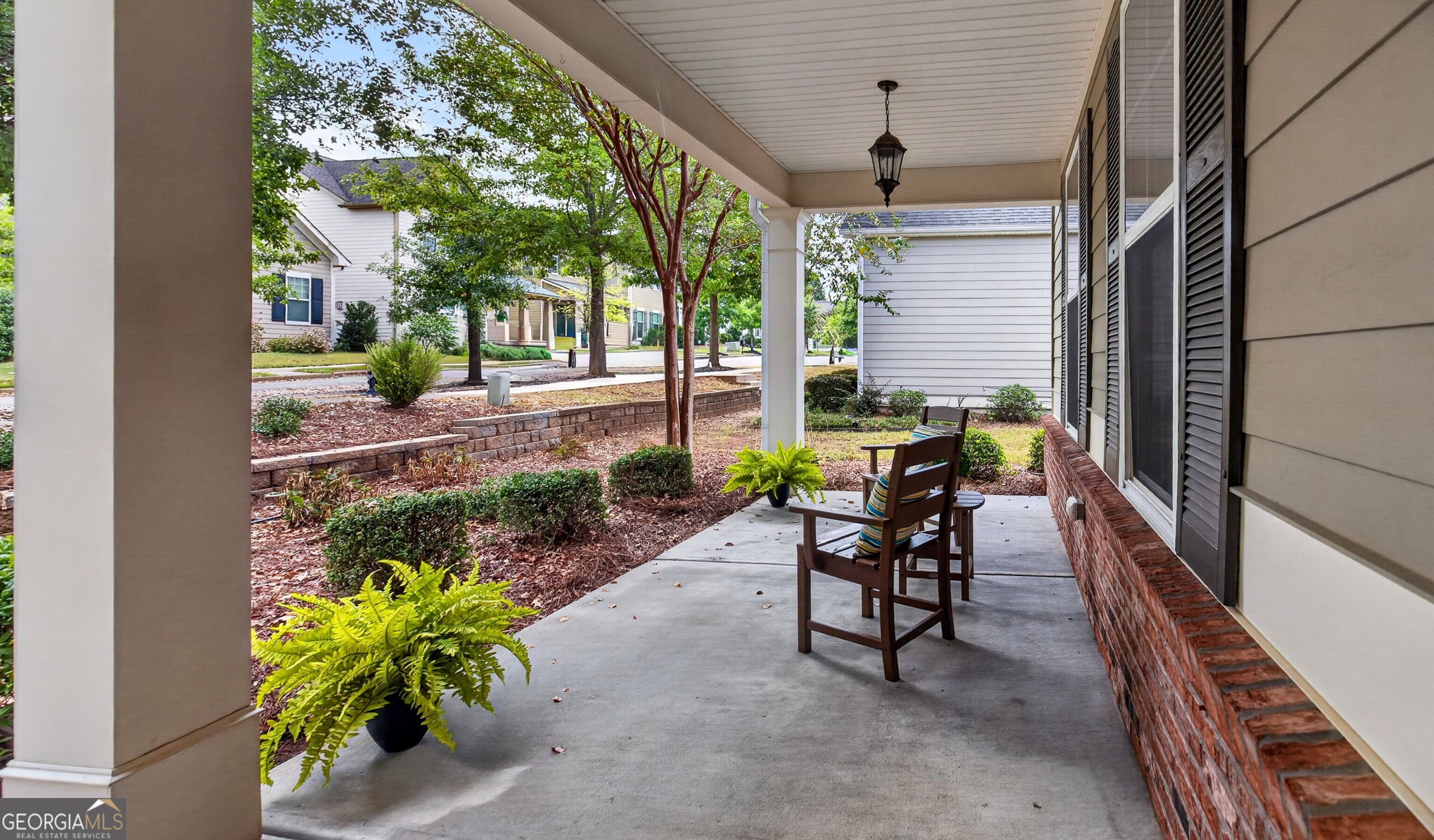Covered front porch with wooden chairs and potted ferns overlooking a landscaped yard.