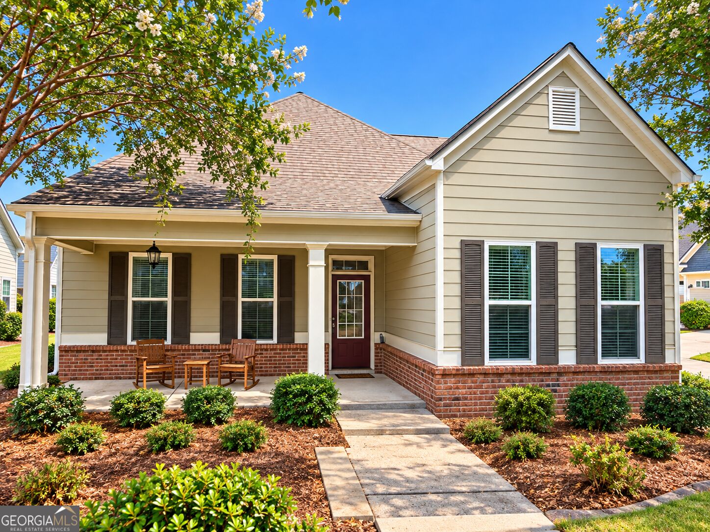 Beige house with a brick base, covered porch, and wooden rocking chairs beneath flowering trees.
