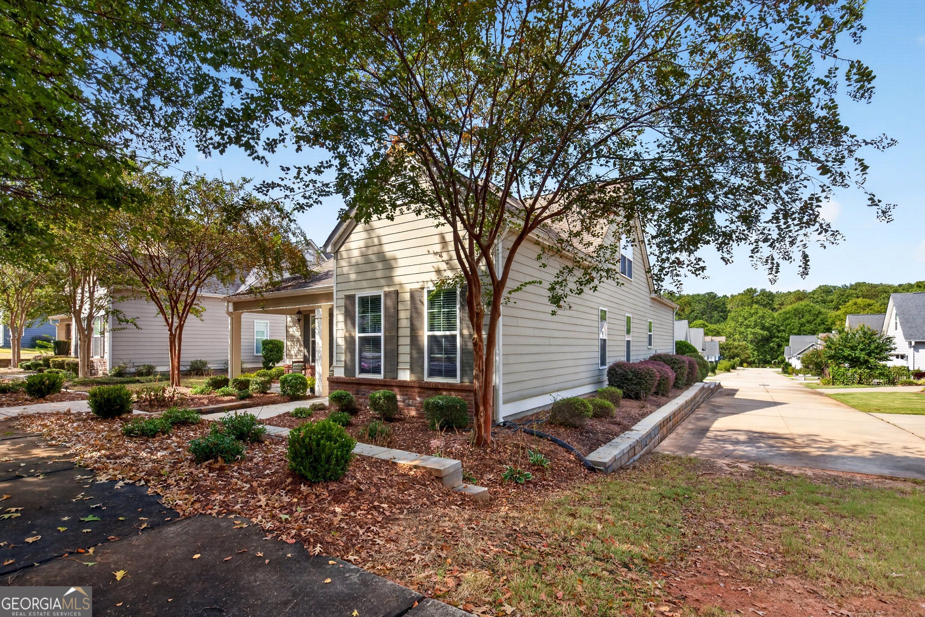 Beige two-story home with a covered entry, mature trees, landscaped beds, and a driveway.