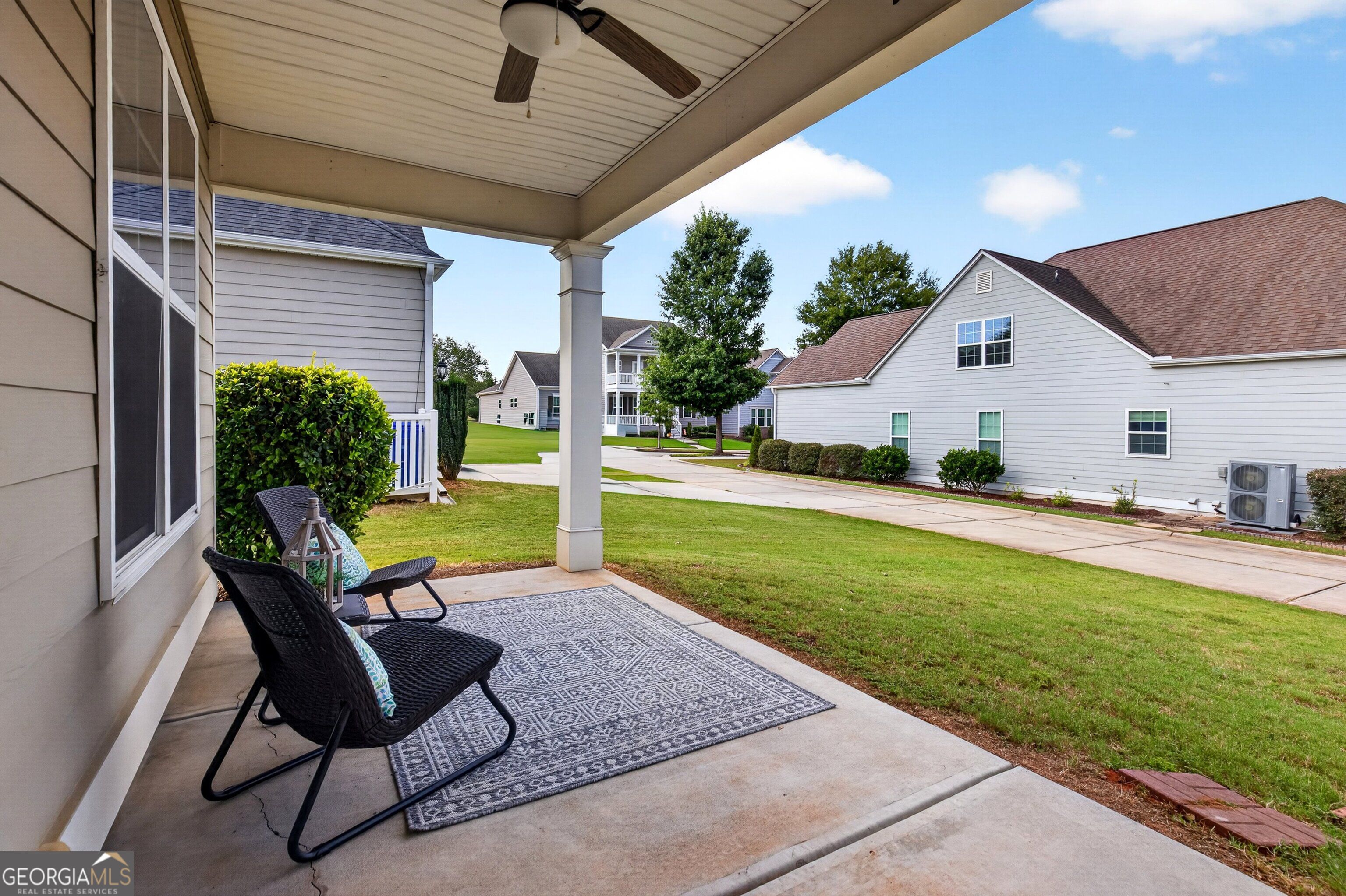 Covered patio with wicker chairs and patterned rug overlooking a lawn and neighboring homes.