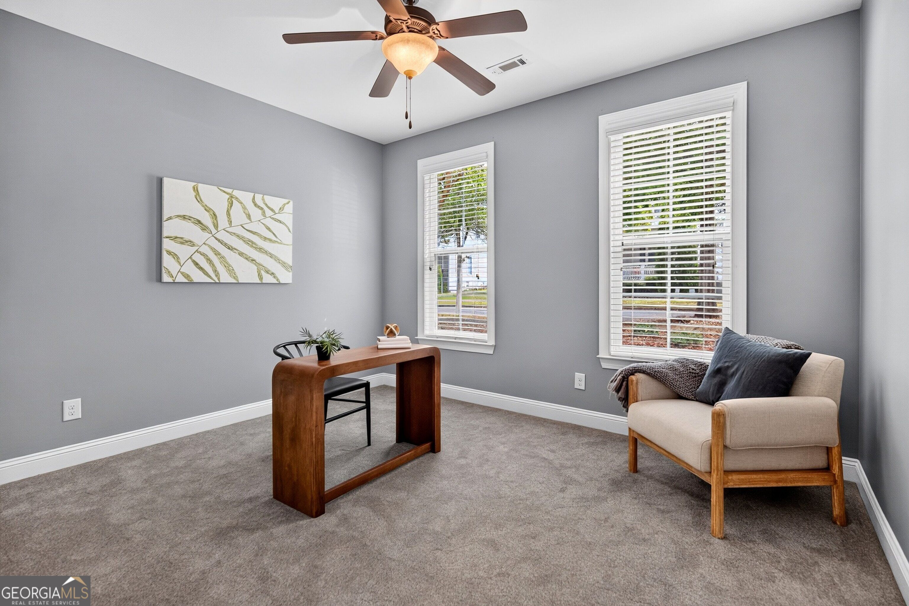 Furnished home office with a wooden desk, cream armchair, and two windows with white blinds.