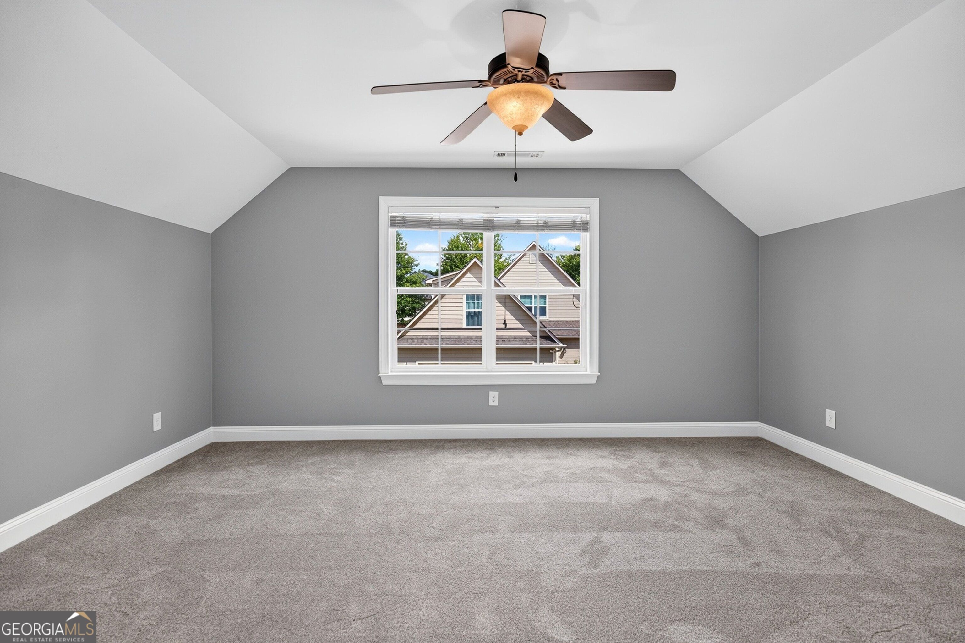 Empty carpeted room with gray walls, vaulted ceiling, ceiling fan, and a window overlooking a neighboring house.