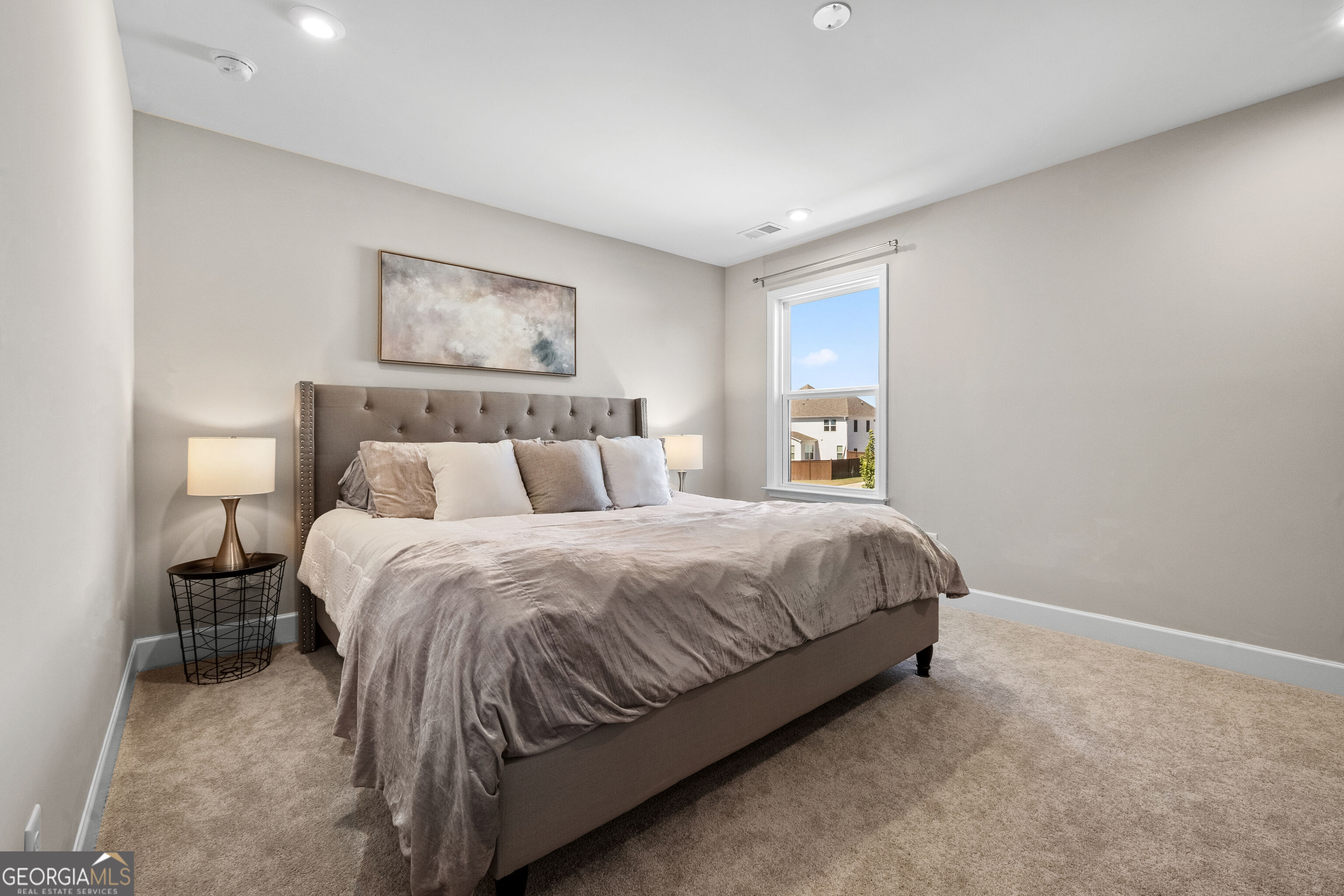 Staged bedroom with a large upholstered bed, tufted headboard, and window overlooking neighboring homes.
