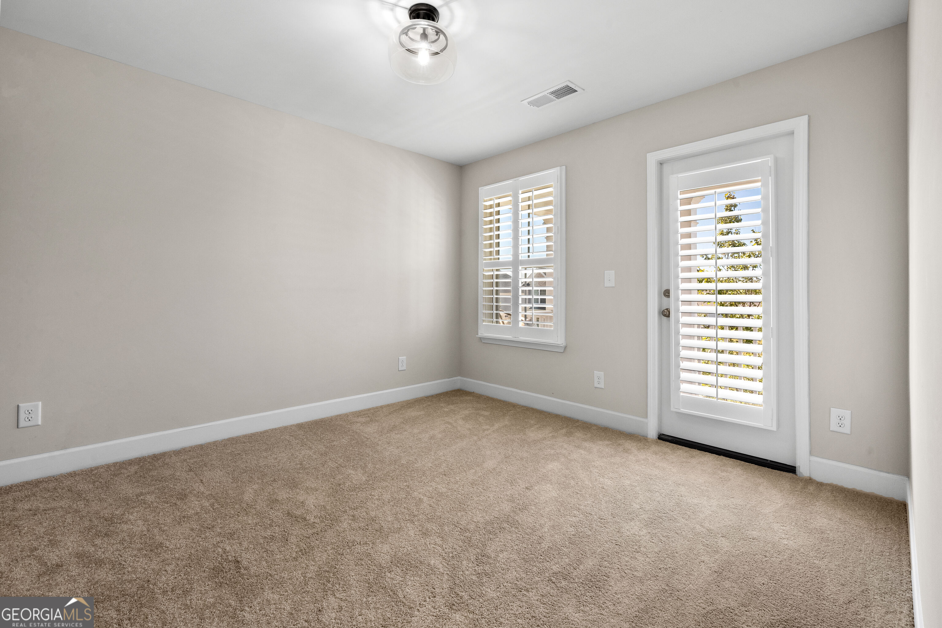 Empty carpeted room with a ceiling fan and plantation shutters on the window and glass door.