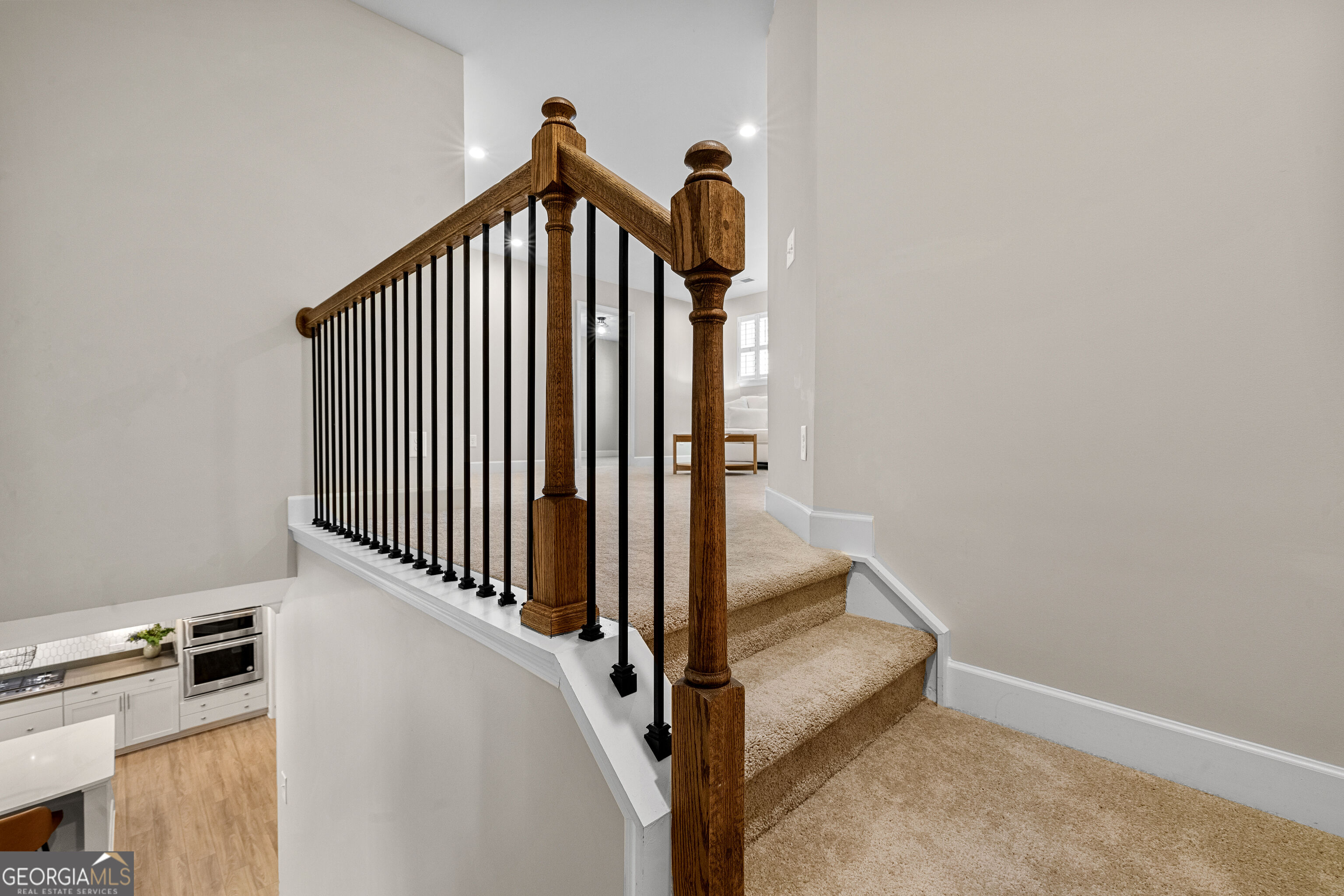 Carpeted staircase with a dark wood handrail and black metal balusters overlooks the kitchen.