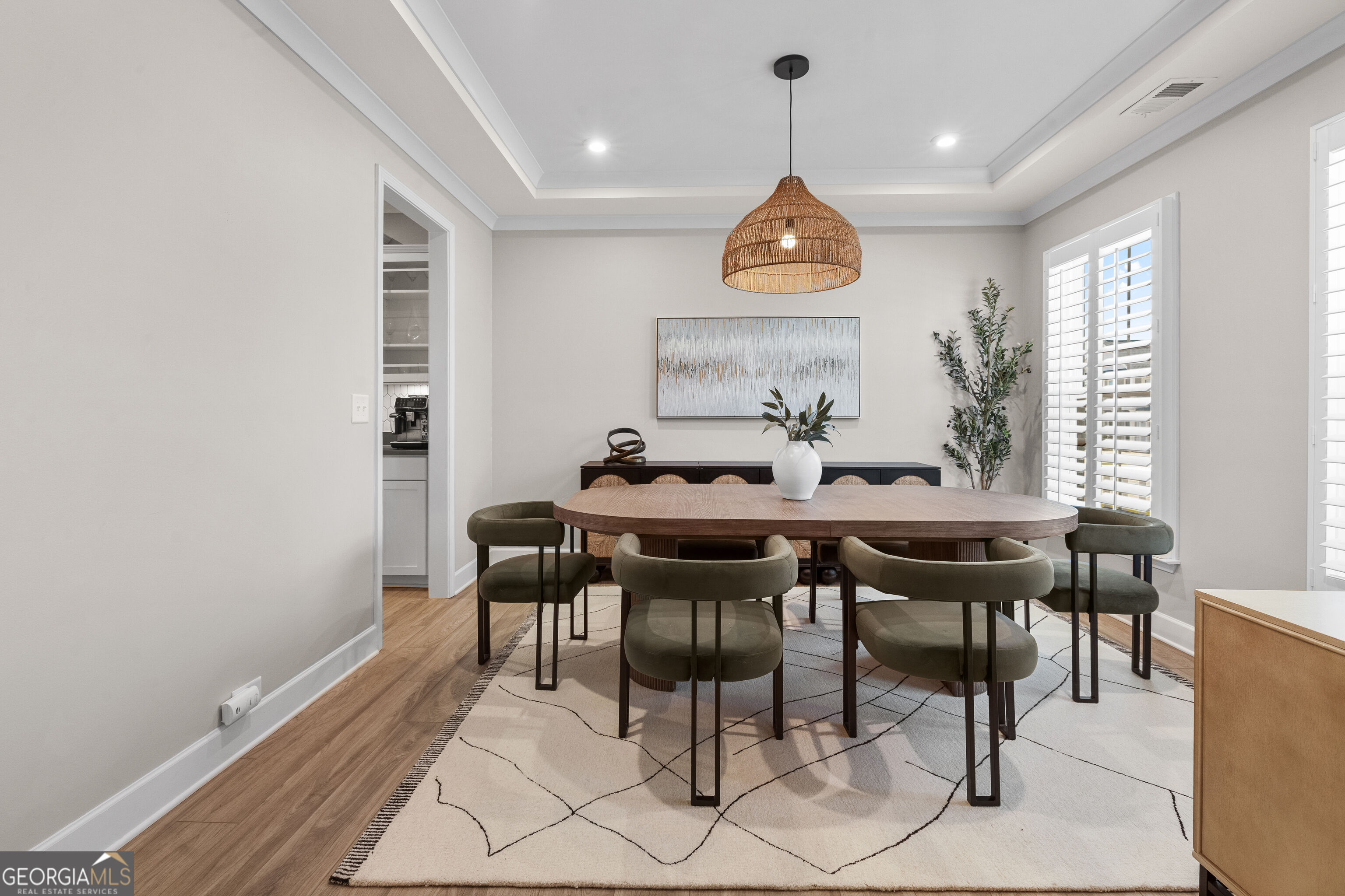 Dining room with a long wood table, olive-green chairs, woven pendant light, and plantation shutters.