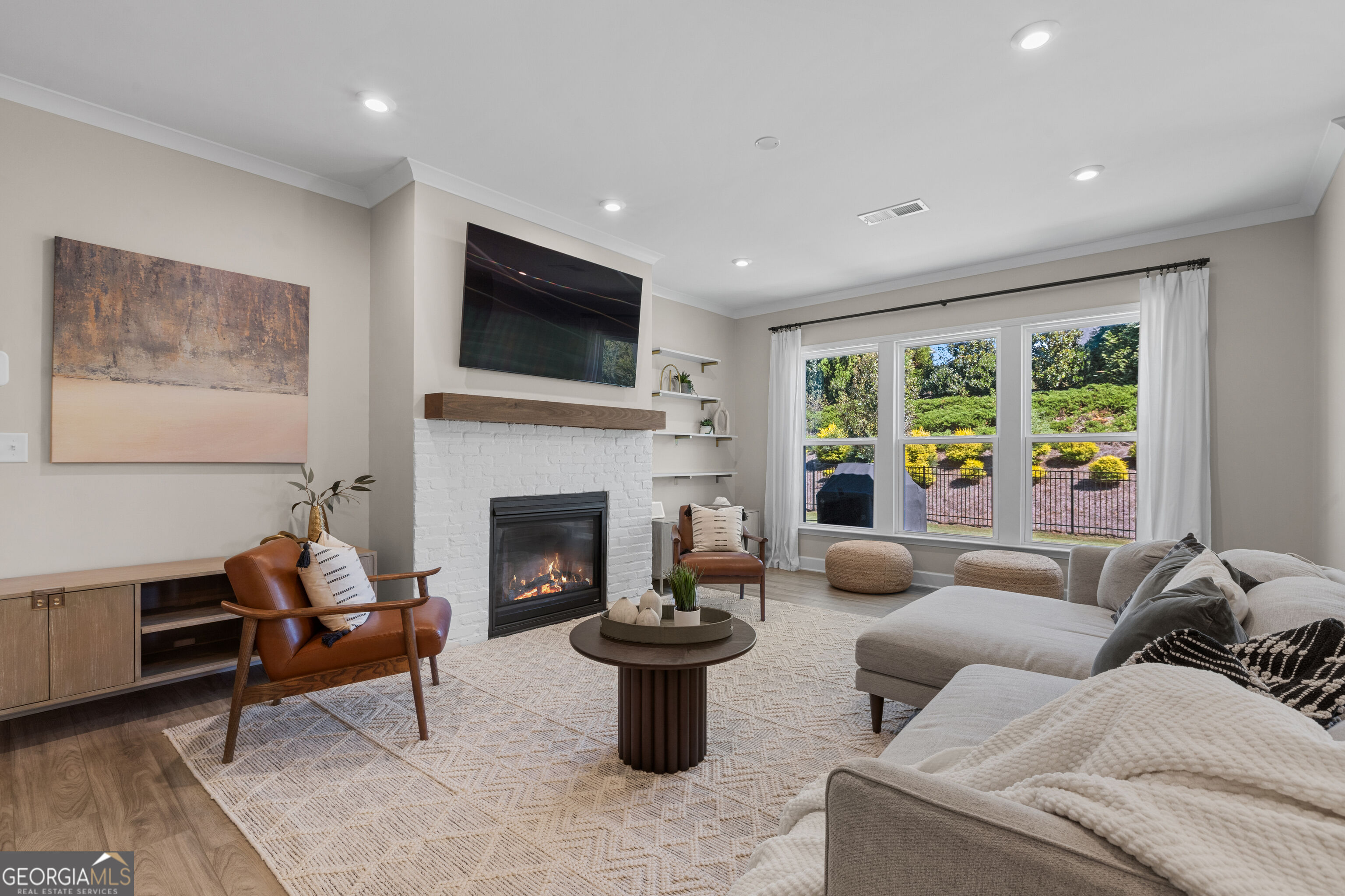 Staged living room with a white brick fireplace, gray sectional, and large windows overlooking the yard.
