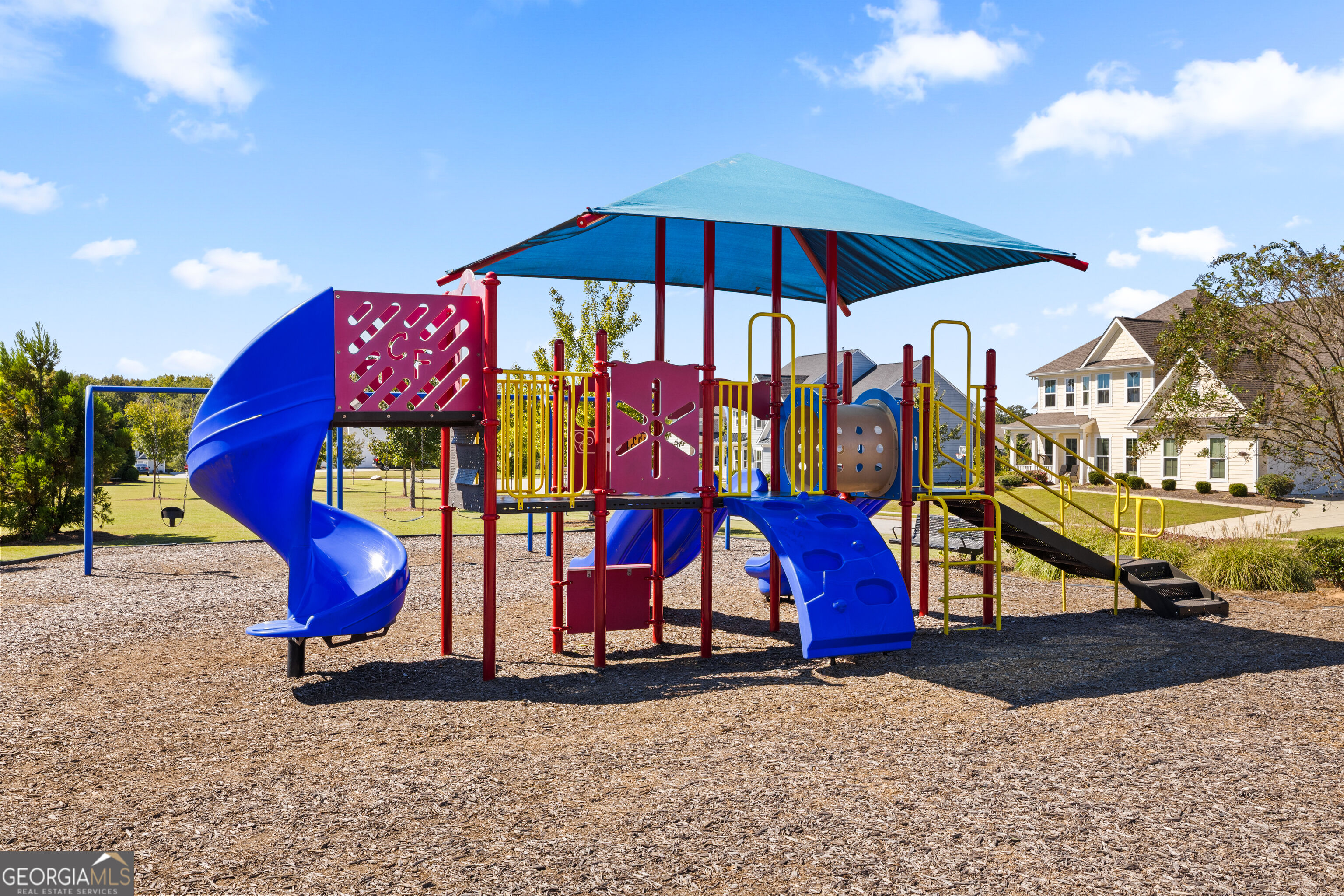 Community playground with blue slides and a shaded climbing structure beside a house.