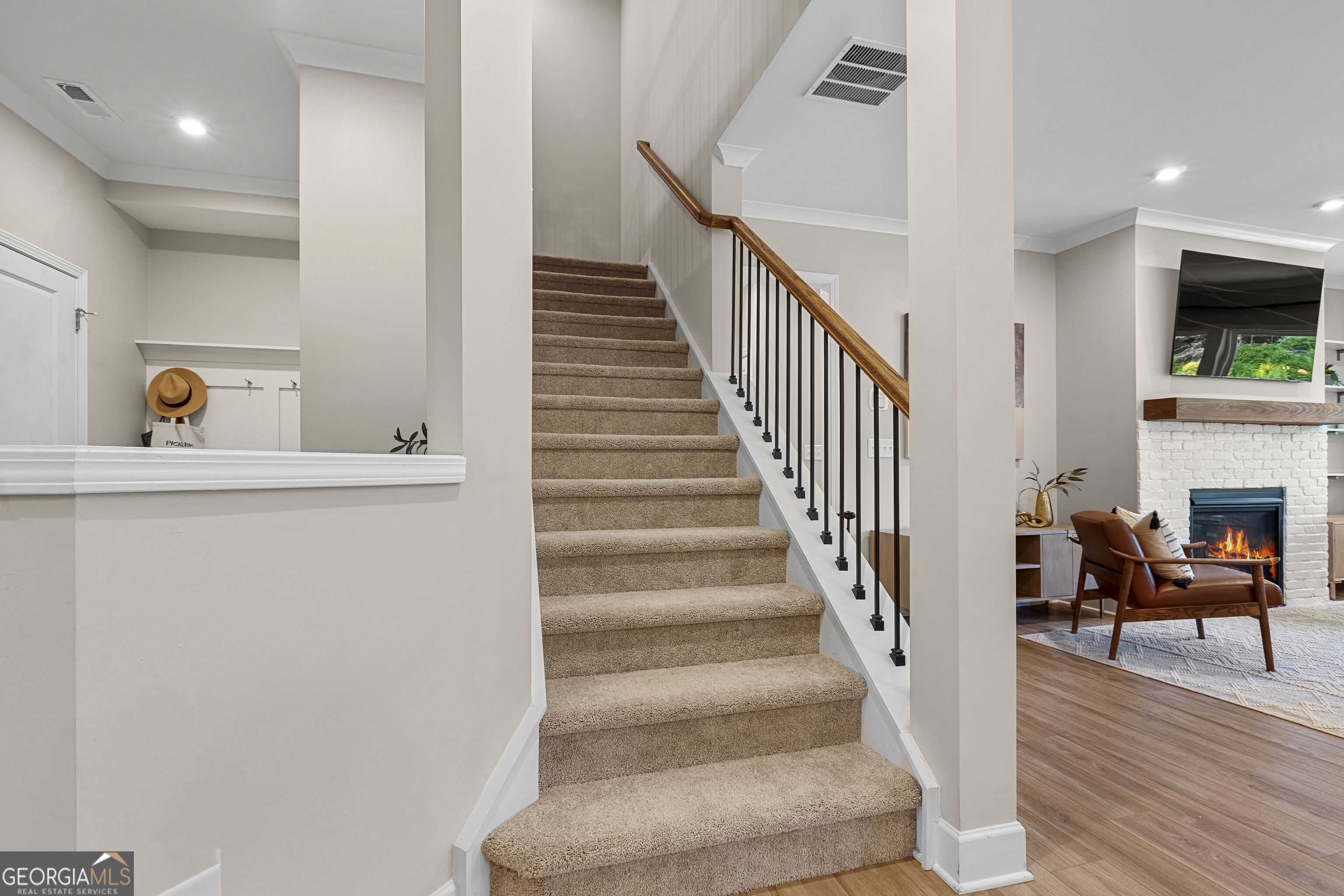 Carpeted staircase with a wood handrail beside a living room with a white brick fireplace.